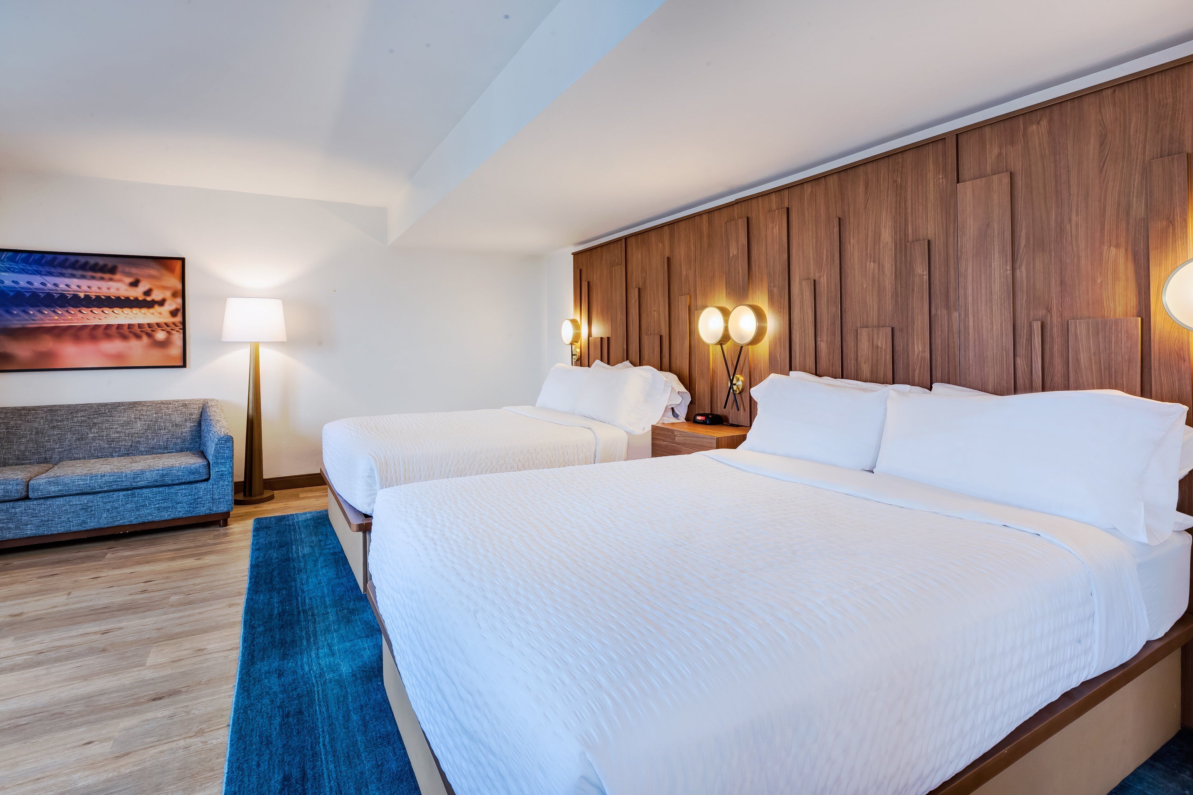 A hotel room with two beds, a sofa, a wooden headboard, and a painting on the wall.