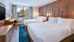 A hotel room with two beds, a desk, a TV, and a large window with a view of buildings.