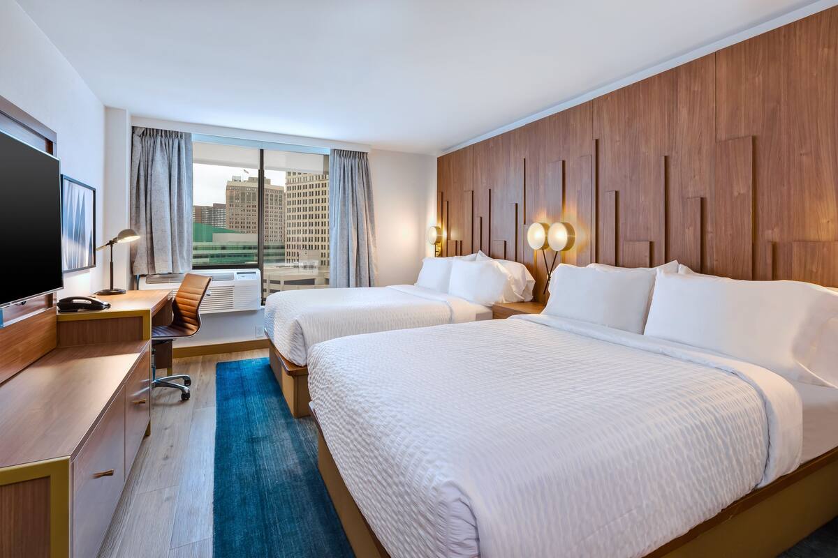A hotel room with two beds, a desk, a TV, and a large window with a view of buildings.