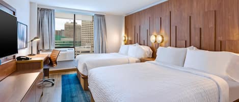 A hotel room with two beds, a desk, a TV, and a large window with a view of buildings.