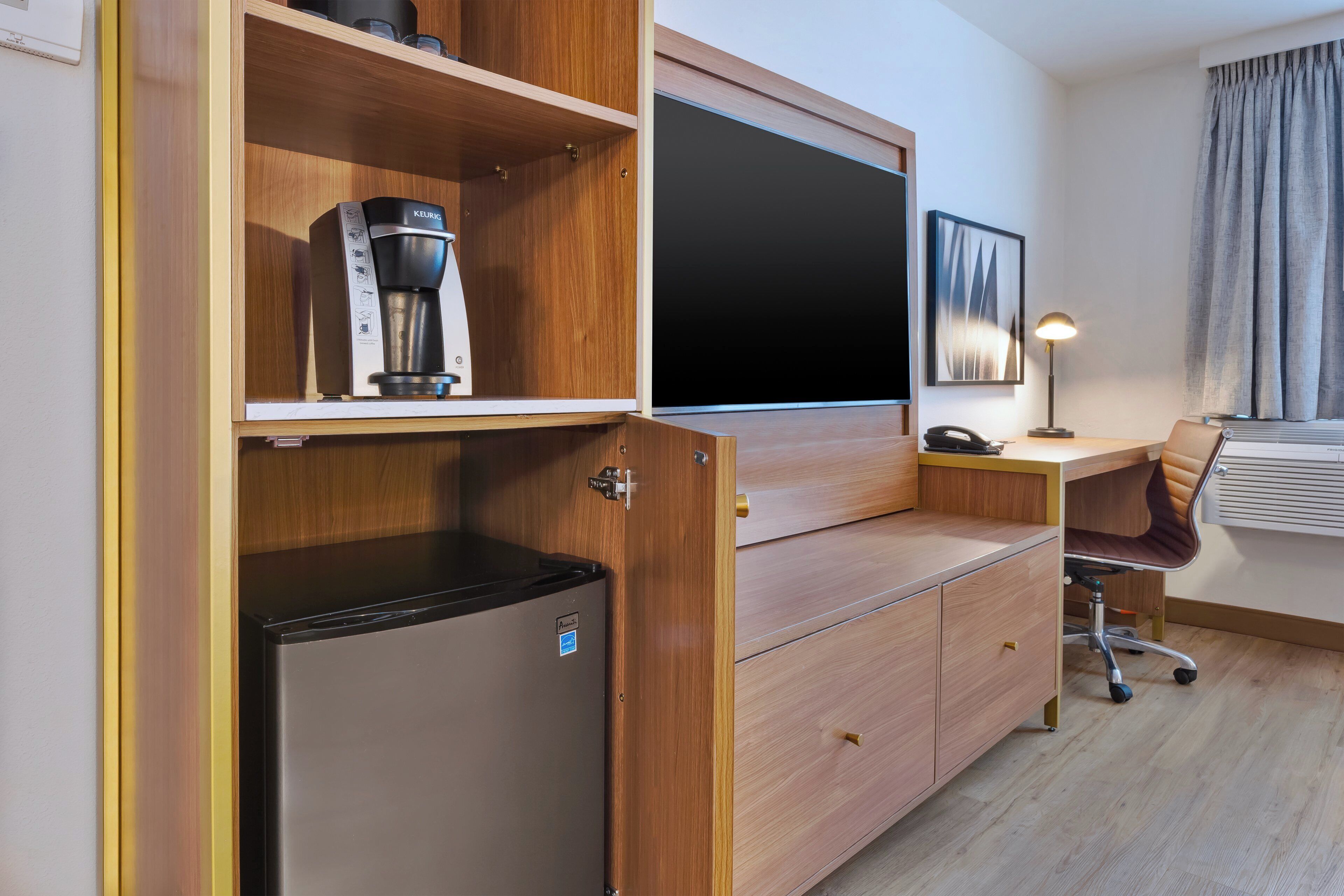 A compact hotel room with a kitchenette, a flat-screen TV, a desk with a lamp, and a chair.