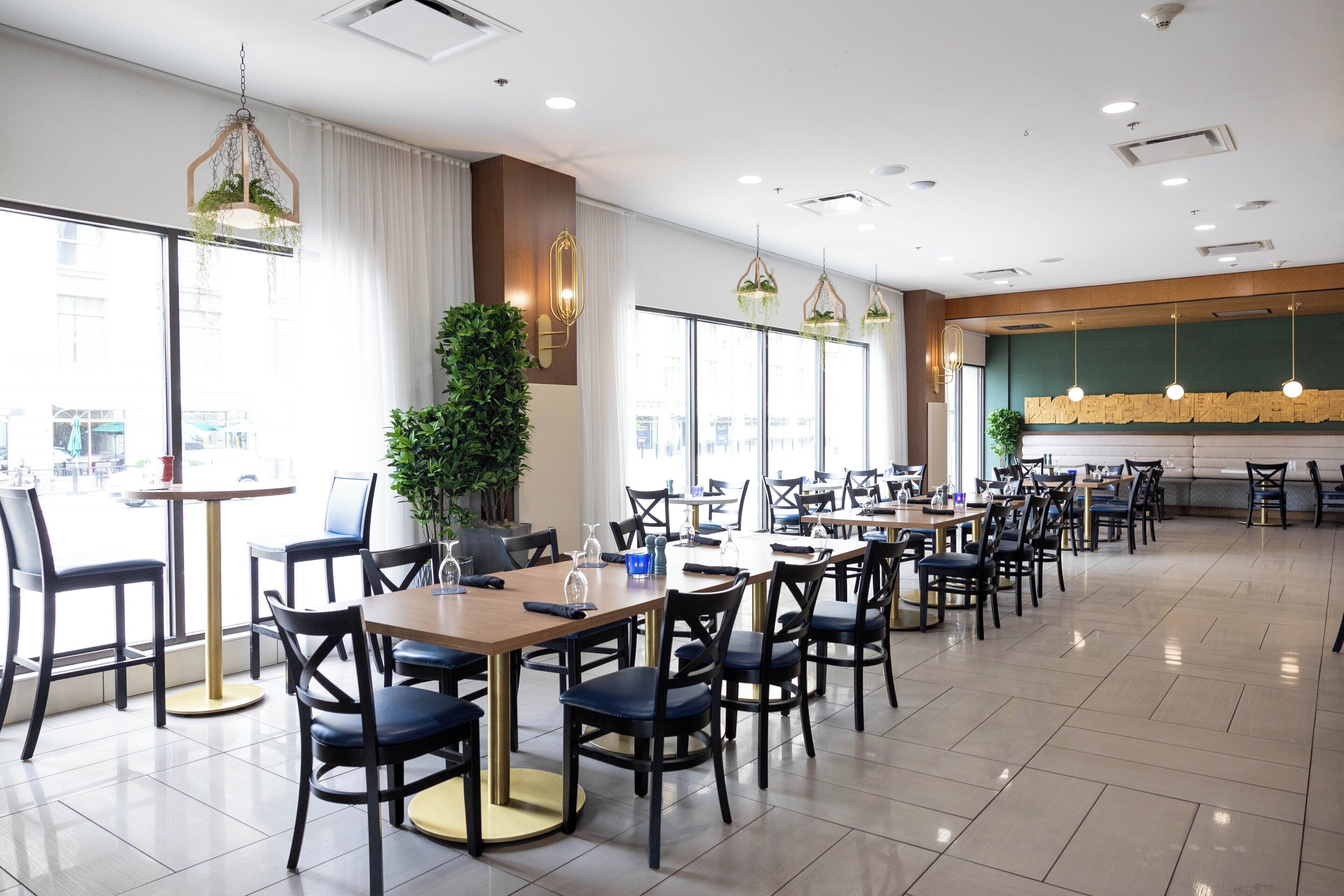 A spacious dining area with multiple tables and chairs, large windows, and hanging plants.