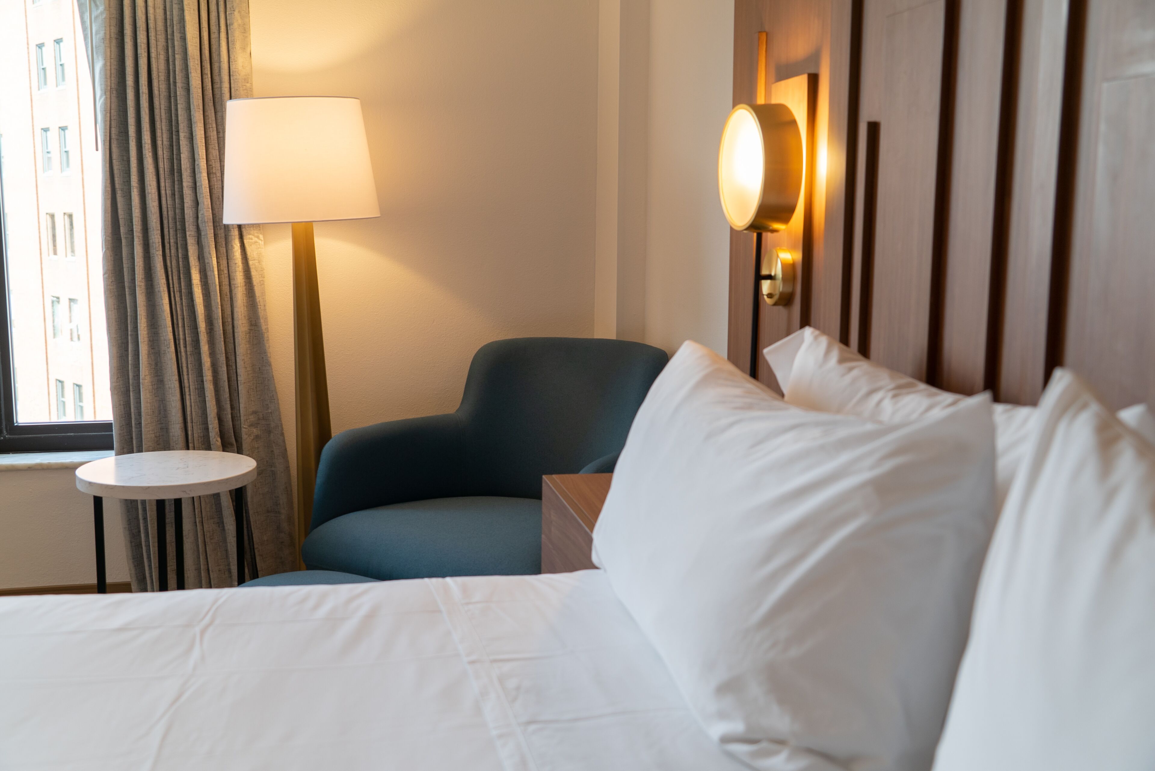 A hotel room with a bed, a chair, a lamp, and a view of a building through a window.