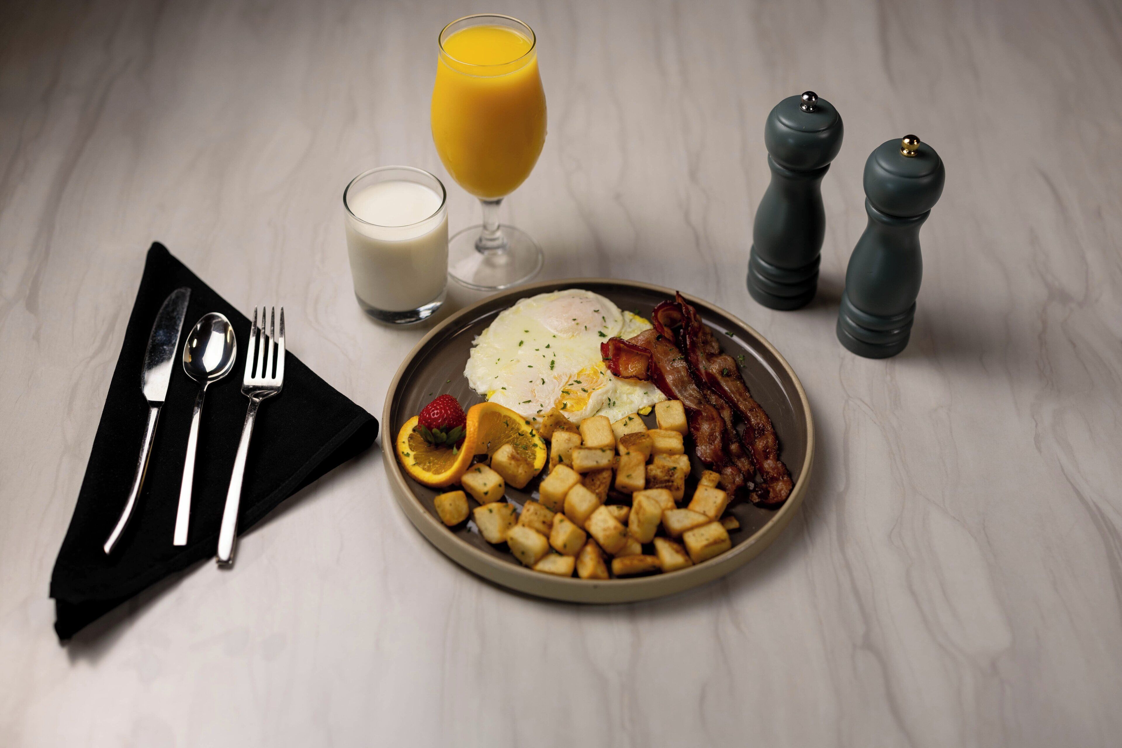 Une assiette de petit-déjeuner comprenant des œufs, du bacon, des toasts et des fruits, accompagnée d’un verre de jus d’orange et d’un verre de lait.
