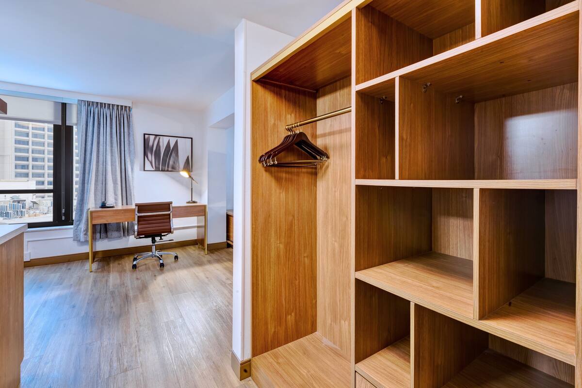 A modern hotel room with a wooden wardrobe, a desk with a chair, and a view of the city through a large window.