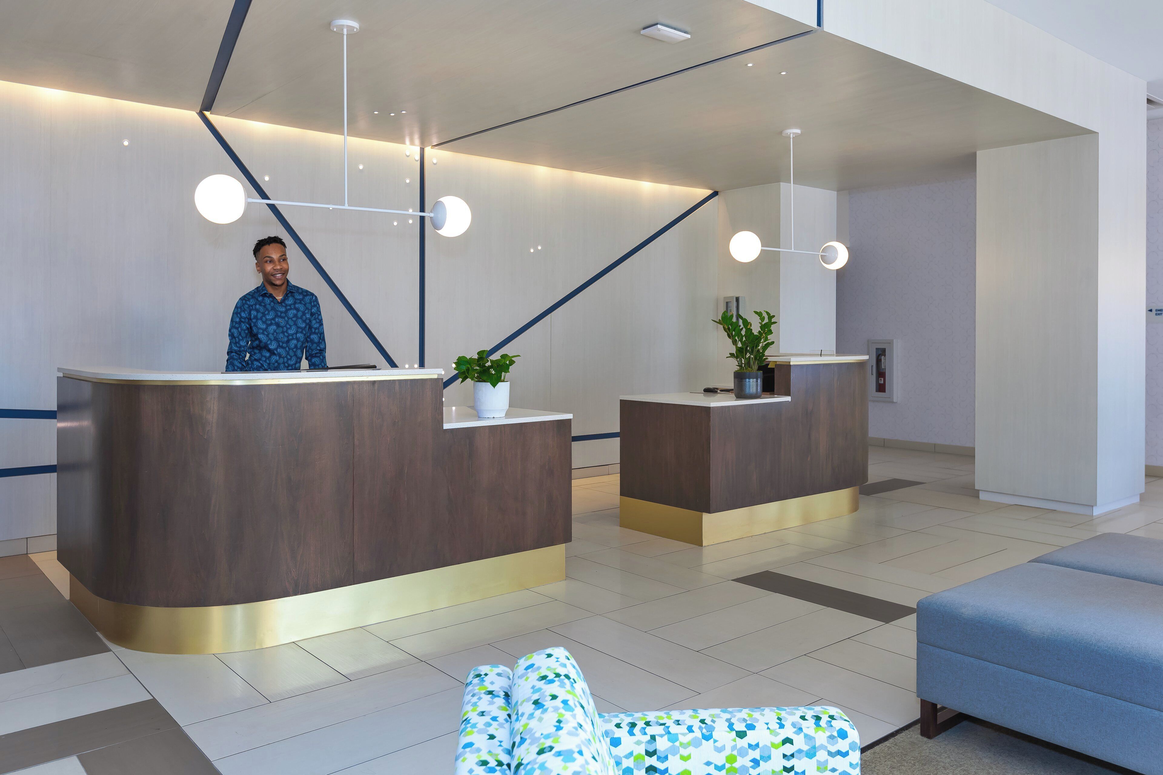 A modern reception desk with a person standing behind it, a potted plant on the desk, and a blue sofa in the foreground.