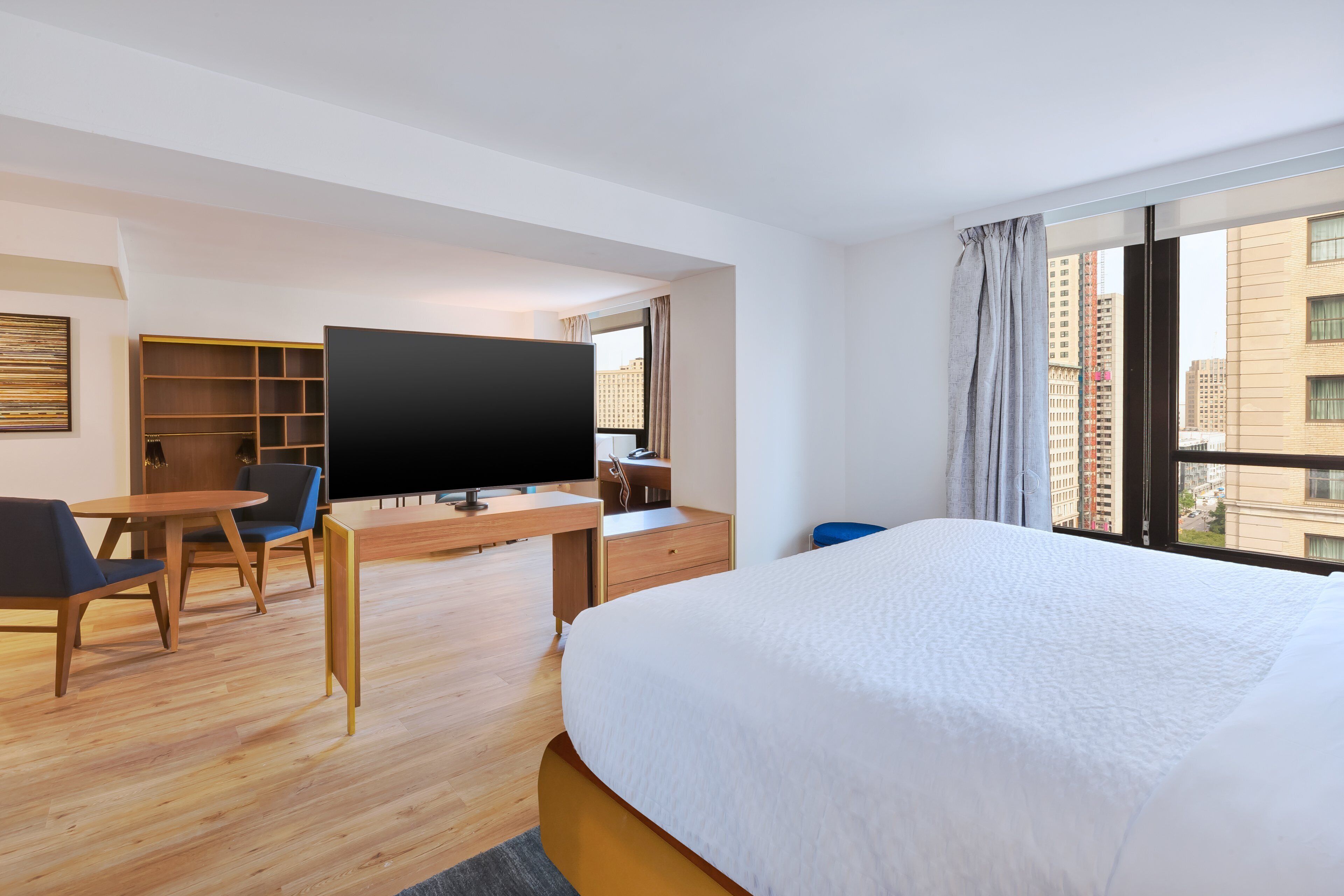 A hotel room with a large bed, a desk with a flat-screen TV, a seating area with chairs, and a view of the city through the window.