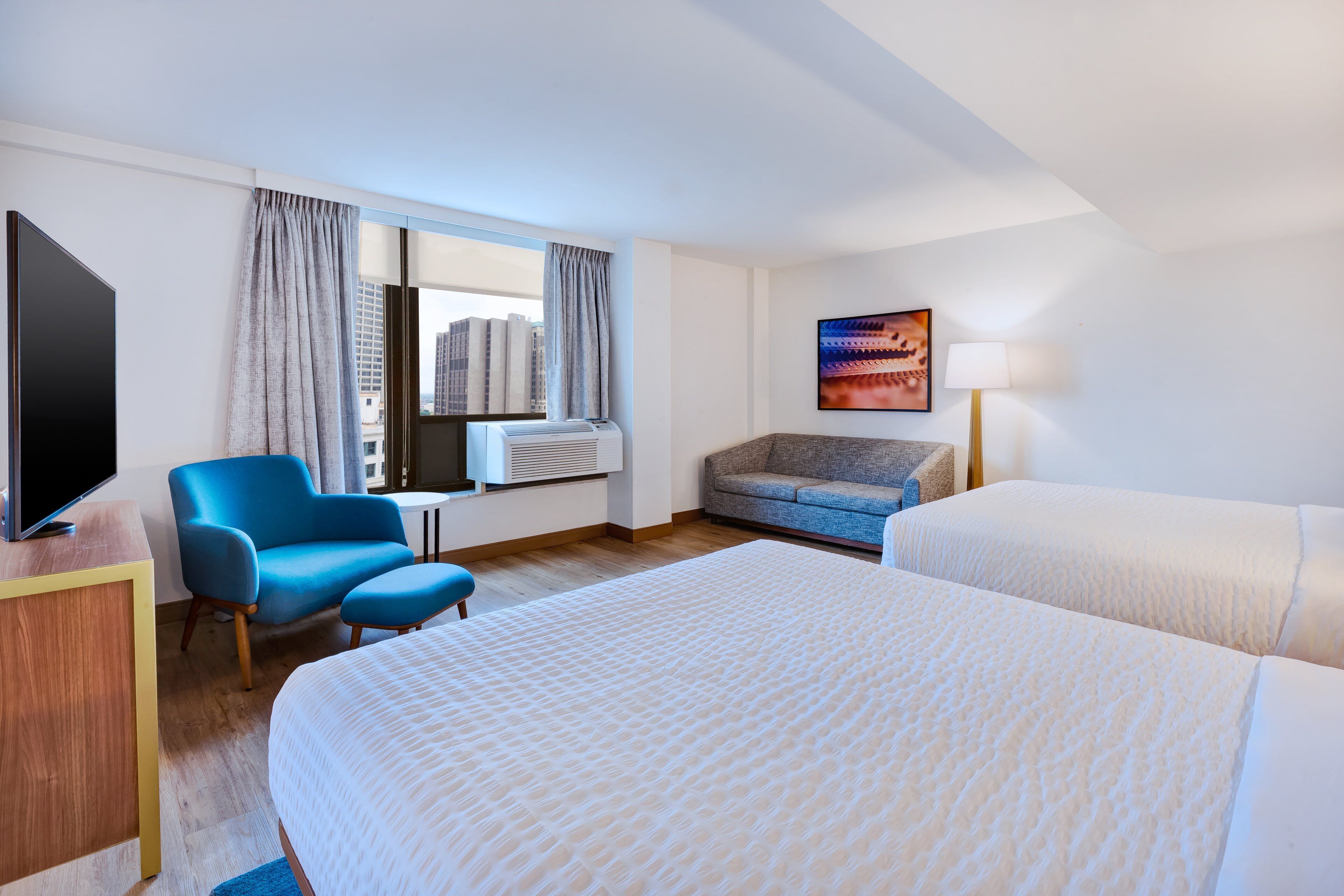 A hotel room with a large bed, a blue armchair, a sofa, a flat-screen TV, an air conditioning unit, and a window with curtains.