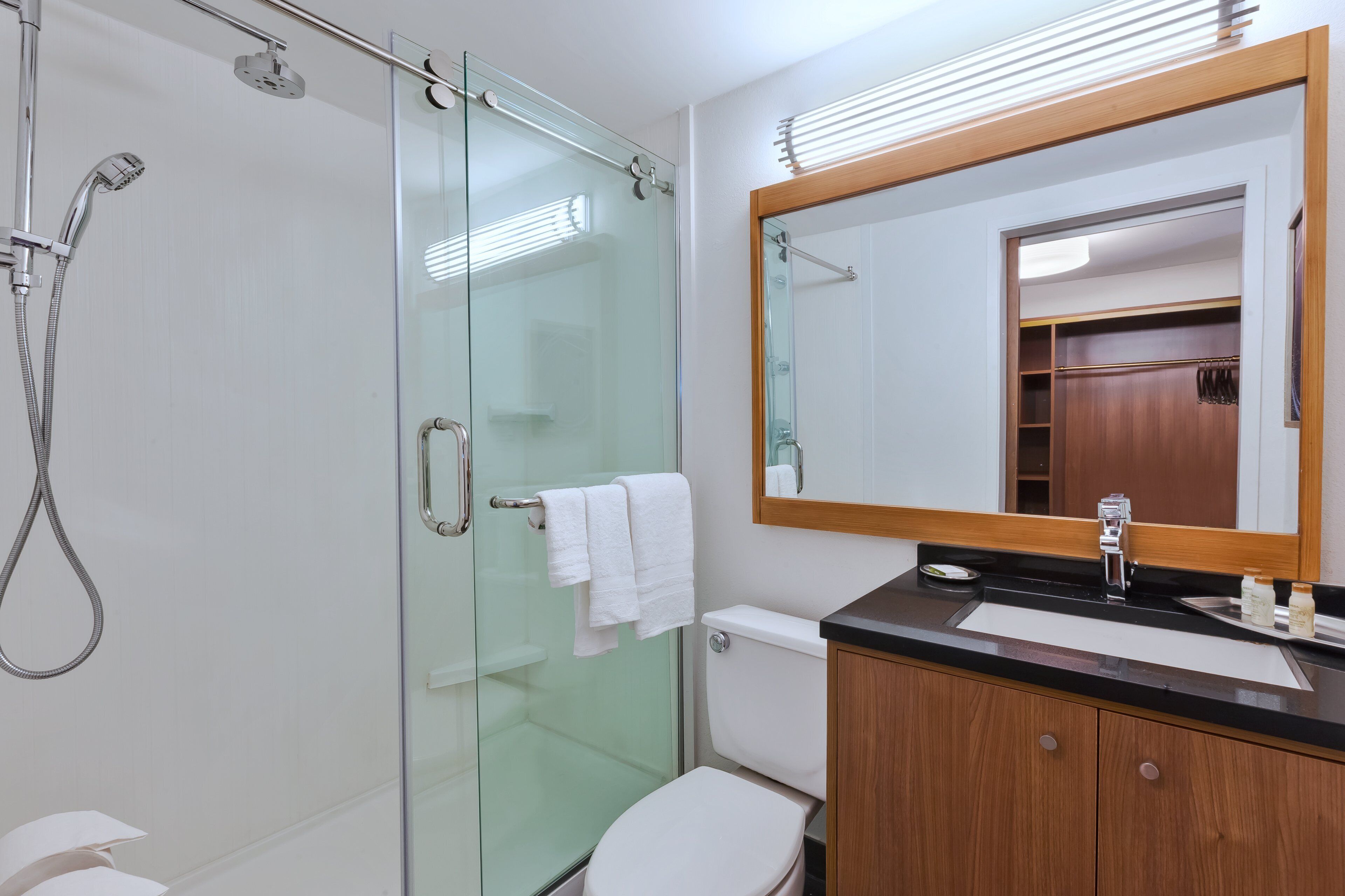 Un baño moderno con ducha envuelta en vidrio, inodoro blanco, tocador de madera con cubierta negra y un espejo grande.