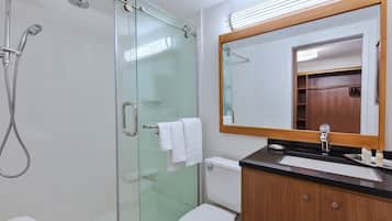 Un baño moderno con ducha envuelta en vidrio, inodoro blanco, tocador de madera con cubierta negra y un espejo grande.