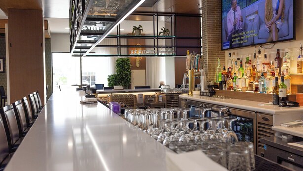 A bar area with a long counter, multiple glasses, and a television screen displaying a show.