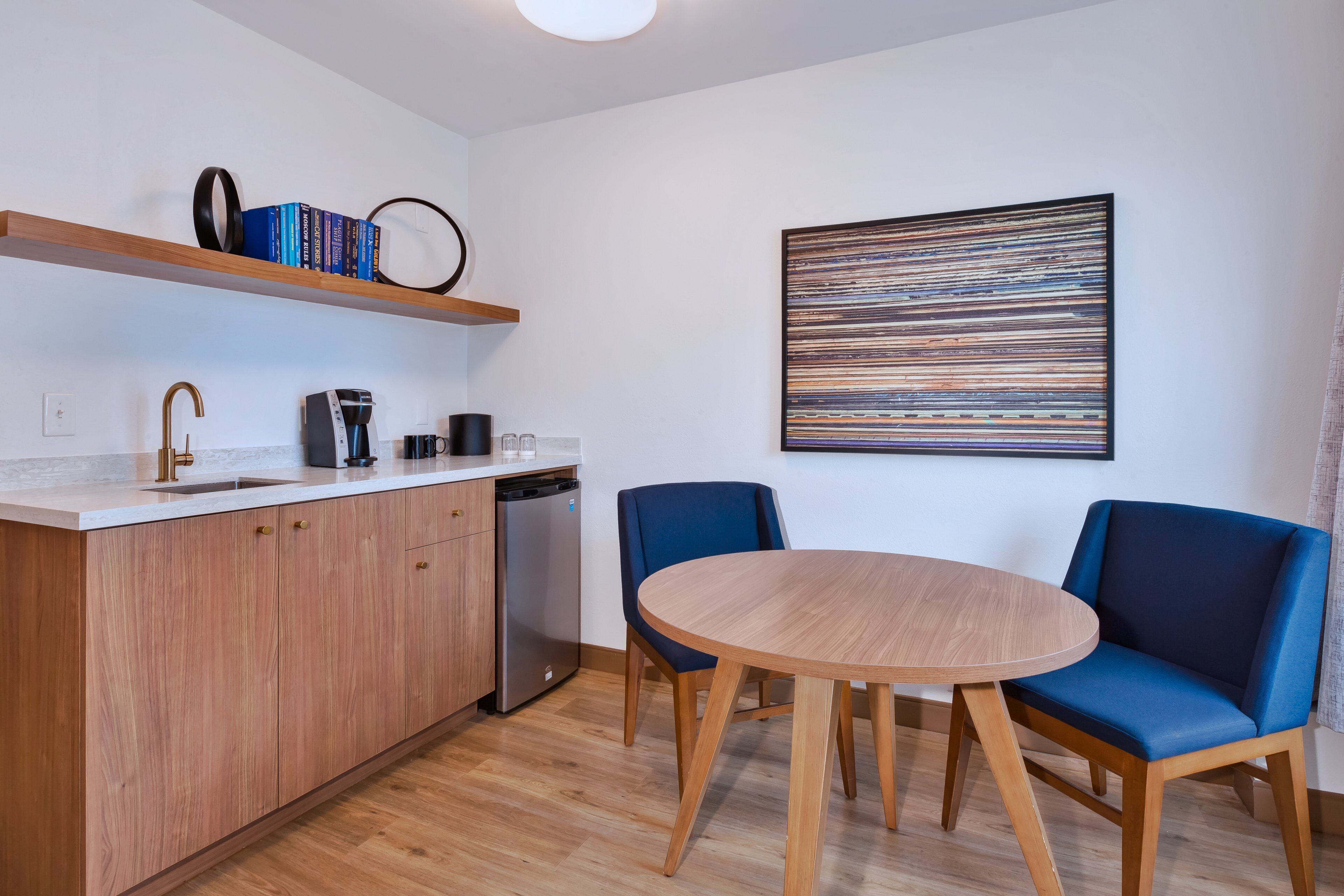 Une cuisine moderne avec des meubles en bois, un lave-vaisselle et une machine à café. On y trouve une table ronde en bois, accompagnée de deux chaises bleues. Un grand tableau abstrait orne le mur.