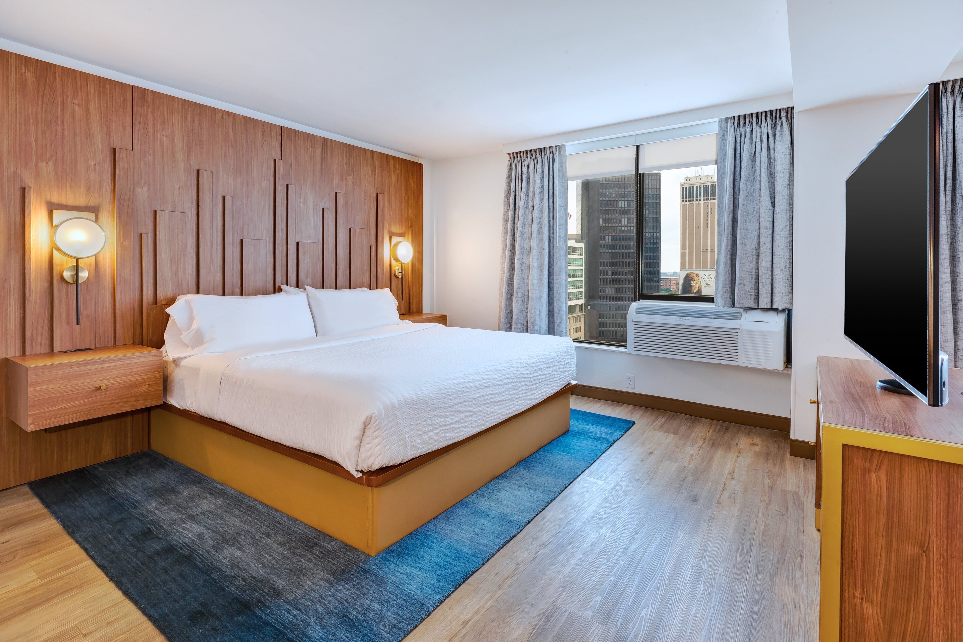 Quarto de hotel moderno com uma cama grande, painel de parede em madeira, duas luminárias de cabeceira, uma janela ampla com cortinas e uma TV de tela plana sobre um móvel de madeira.