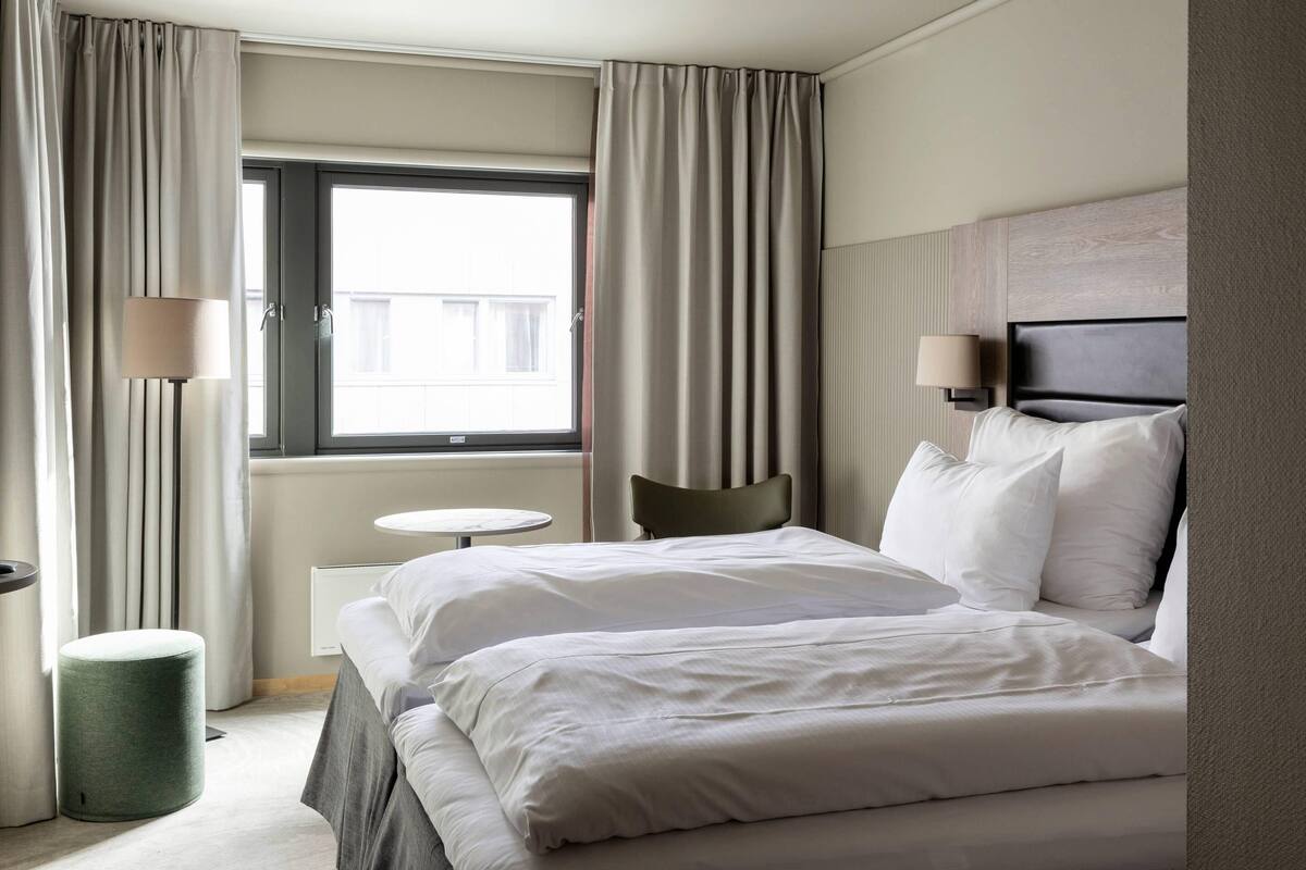A hotel room with a bed, a chair, a small table, a lamp, and a window with curtains.