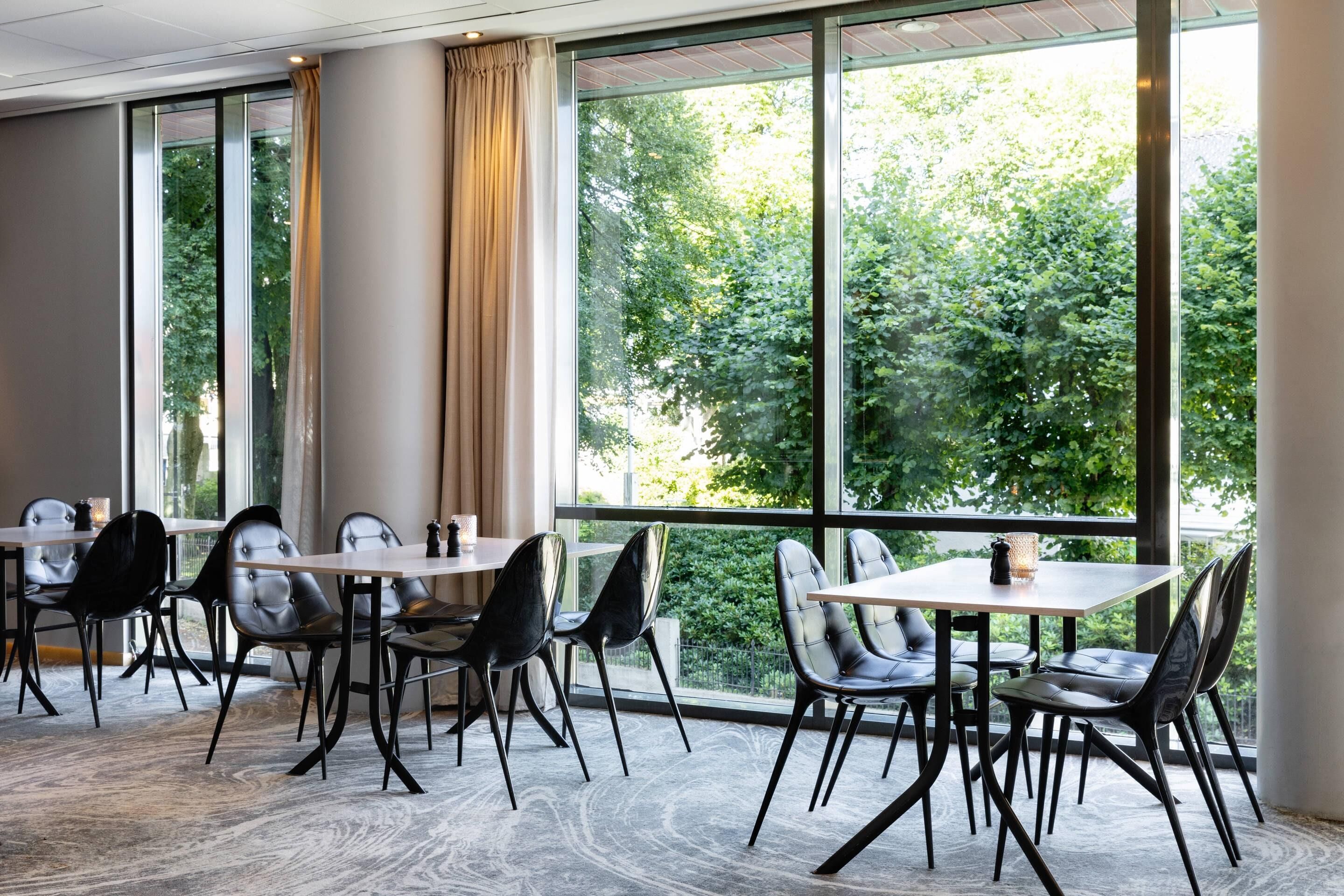 A modern dining area with tables and chairs, large windows with a view of greenery, and a patterned carpet.
