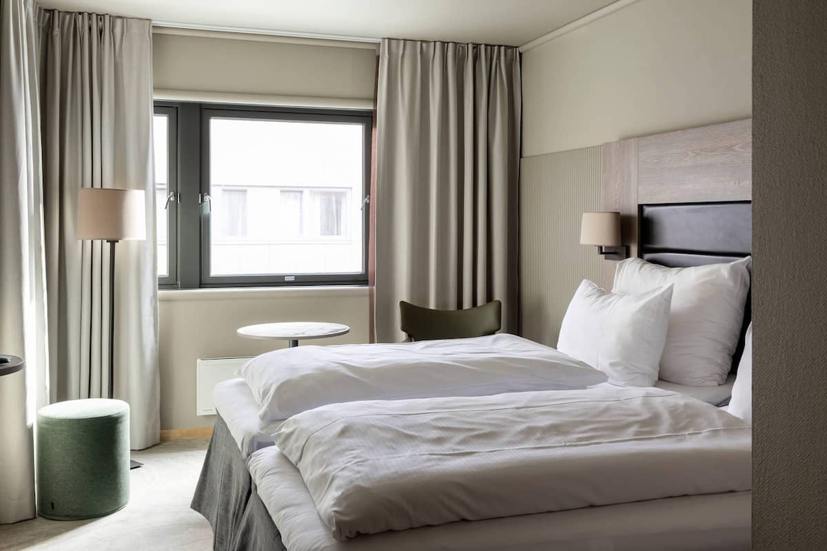 A hotel room with a bed, a chair, a small table, a lamp, and a window with curtains.
