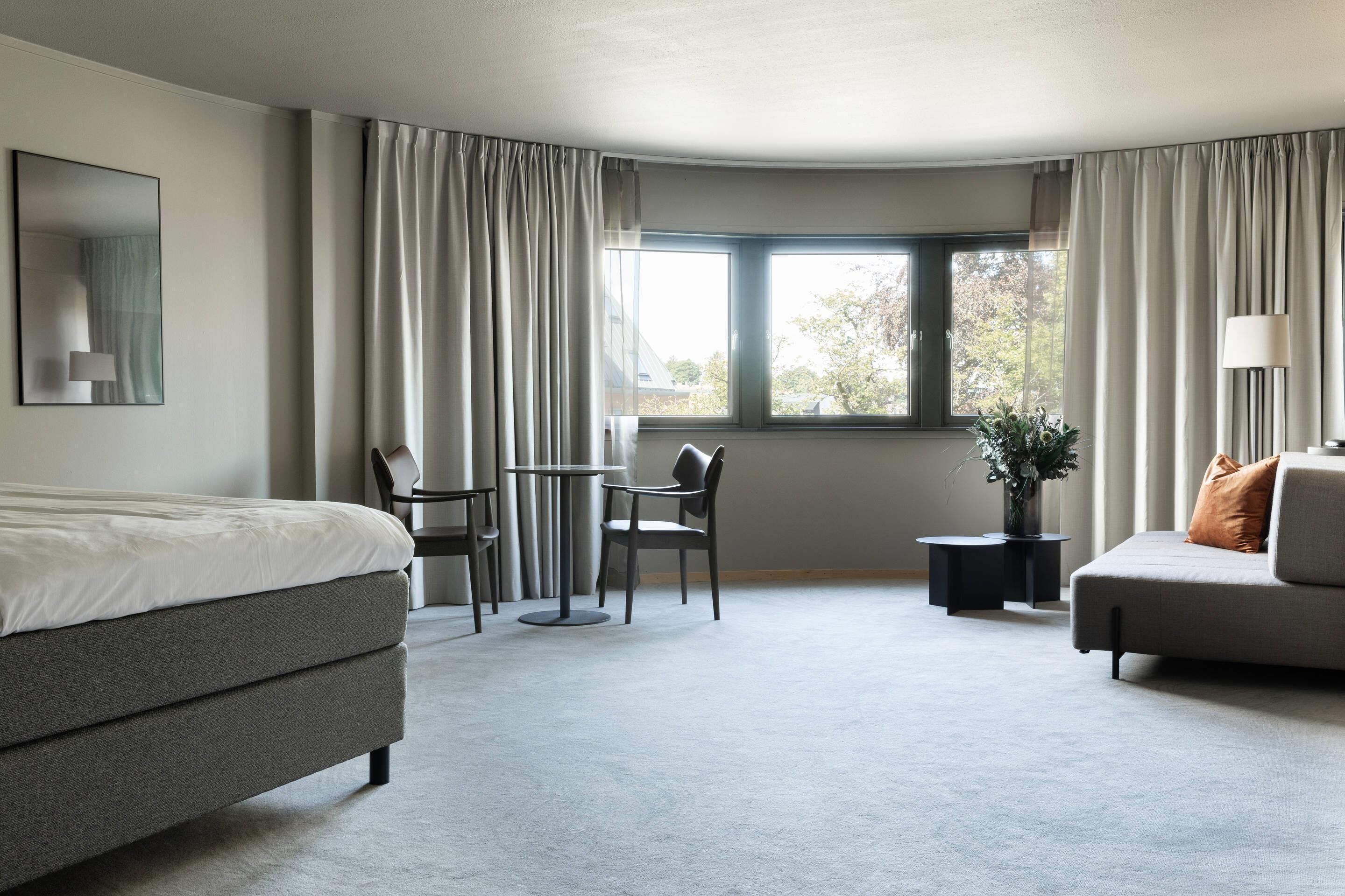 A modern hotel room with a bed, a sofa, a dining table with chairs, a small side table with a plant, and a window with curtains.