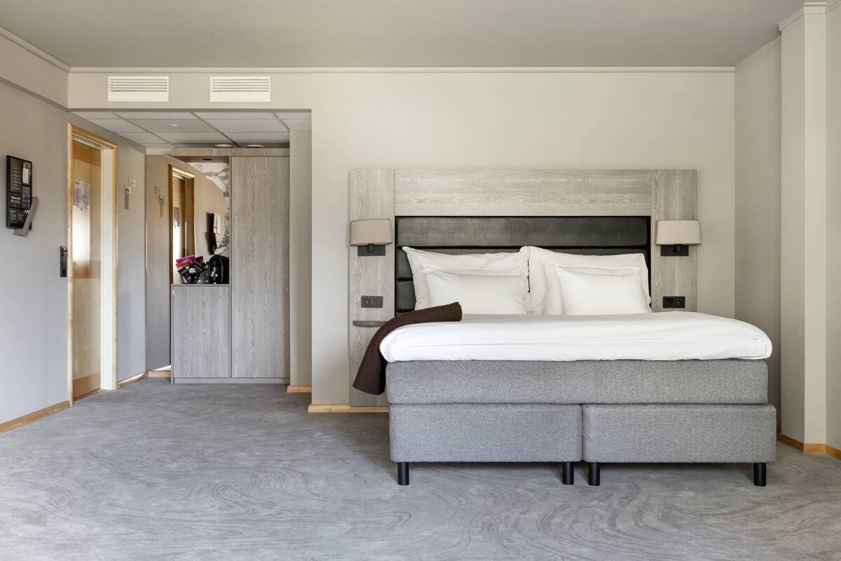 A modern hotel room with a large bed, a headboard, two bedside lamps, and a carpeted floor.