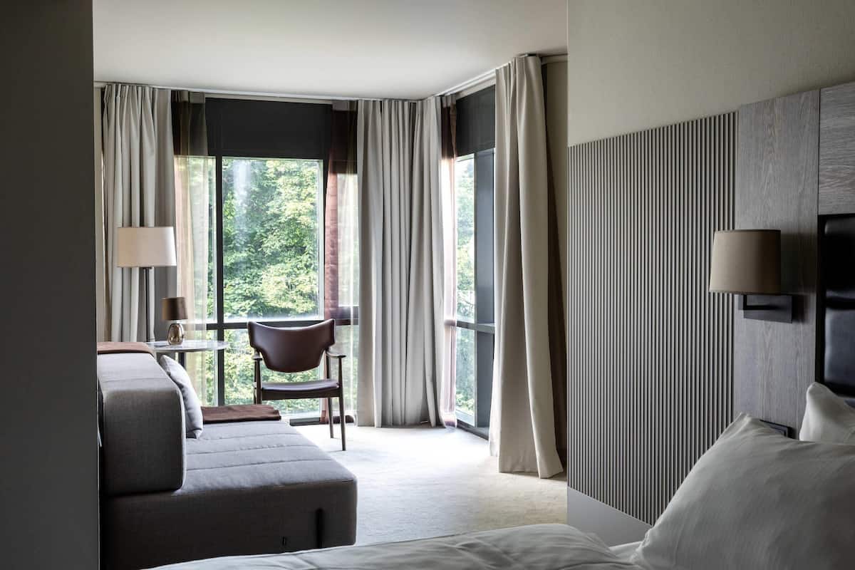 A hotel room with a bed, a chair, a desk, and a large window with curtains.