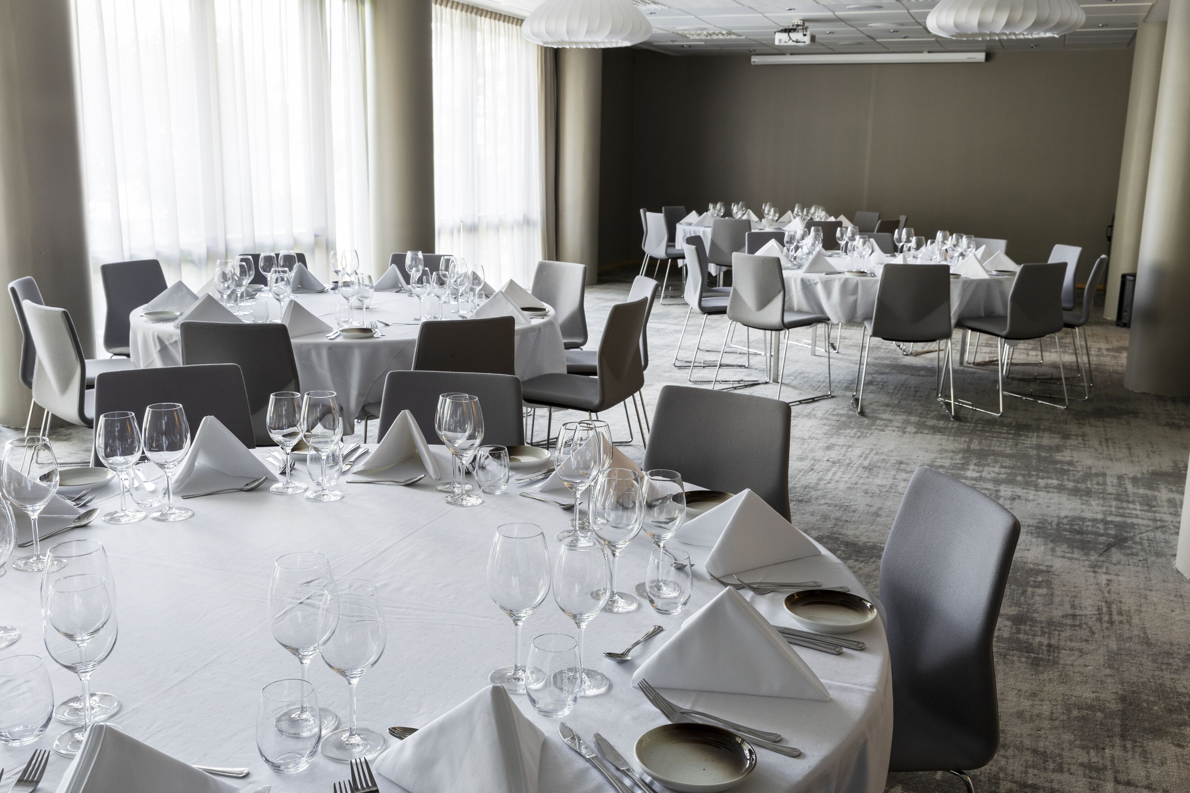 A conference room with round tables set for dining, equipped with chairs, plates, cutlery, and wine glasses.