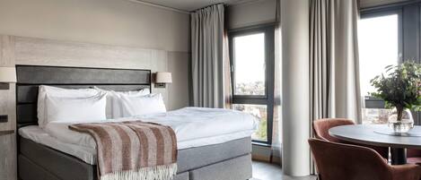 A modern hotel room with a large bed, a dining table, and a view of the city.