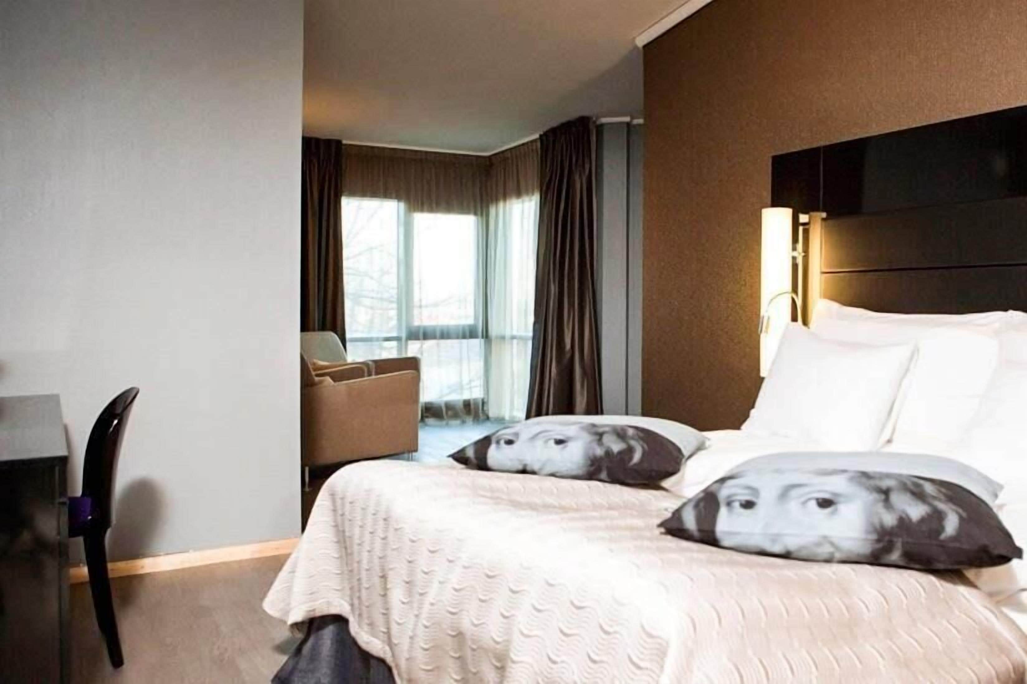 A hotel room with a bed, a chair, a desk, and a window with curtains.