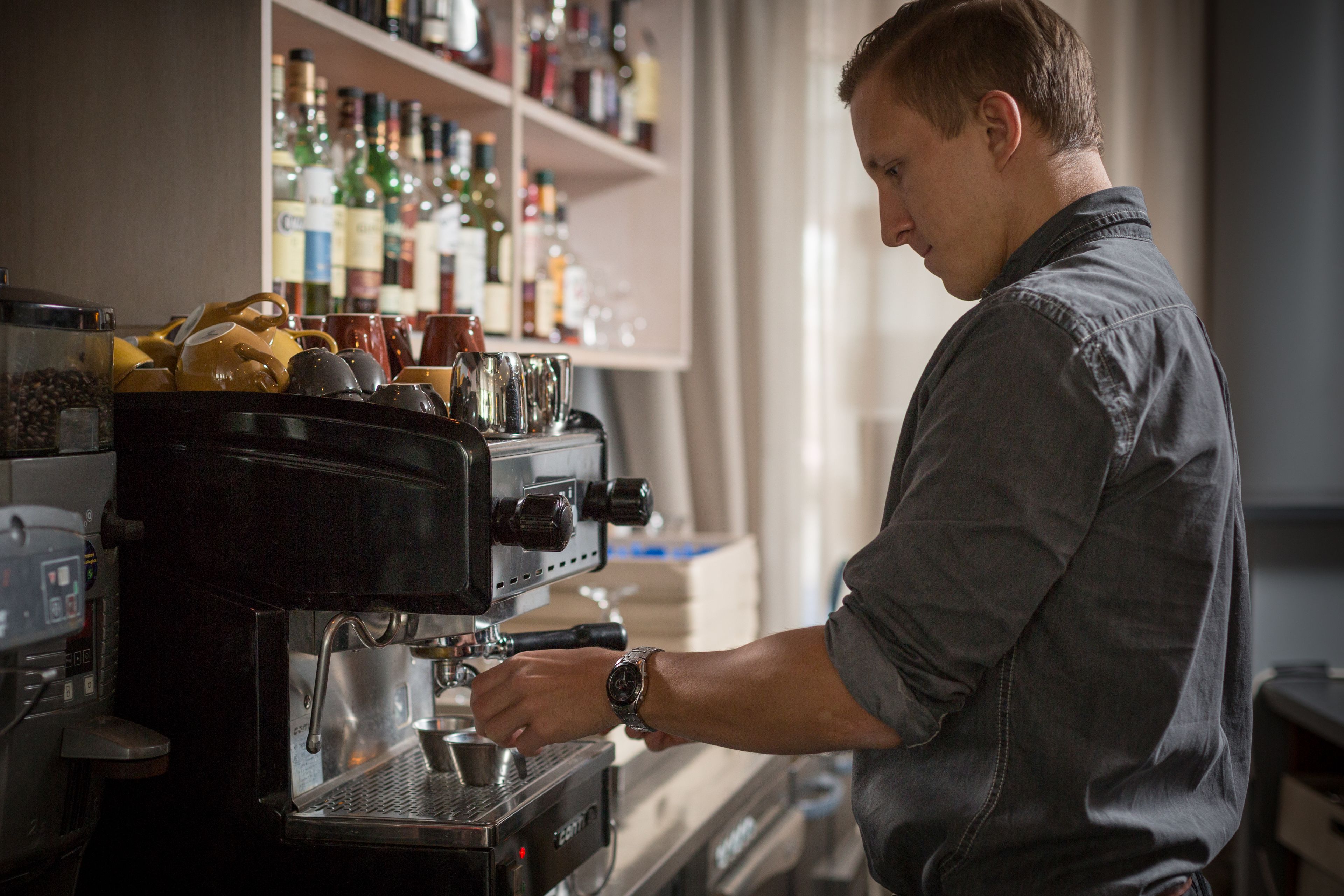 A barista at a coffee machine in a cafe with shelves of various bottles in the background.