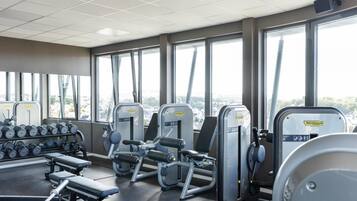 A gym with weight machines, dumbbells, and benches, large windows with a city view.
