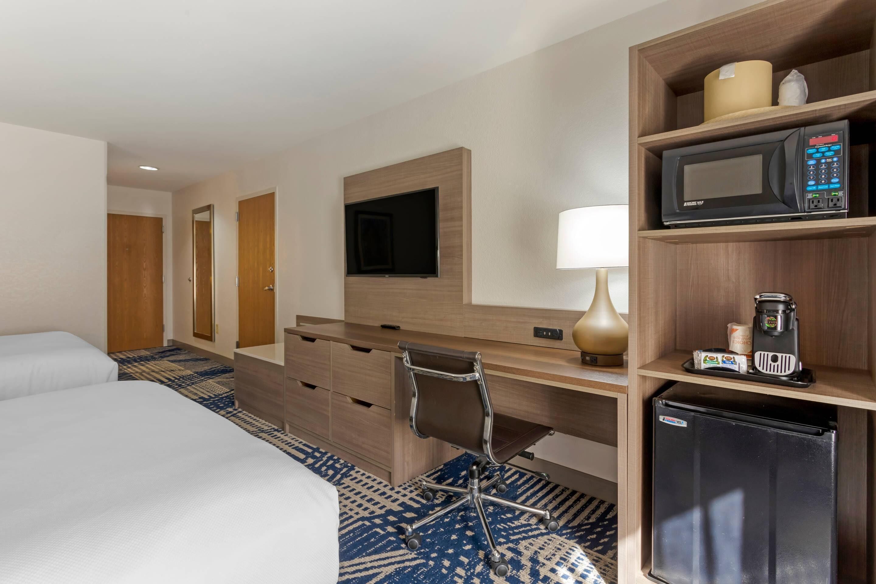 A hotel room with a bed, a desk with a chair, a television, a microwave, a refrigerator, and a coffee maker.