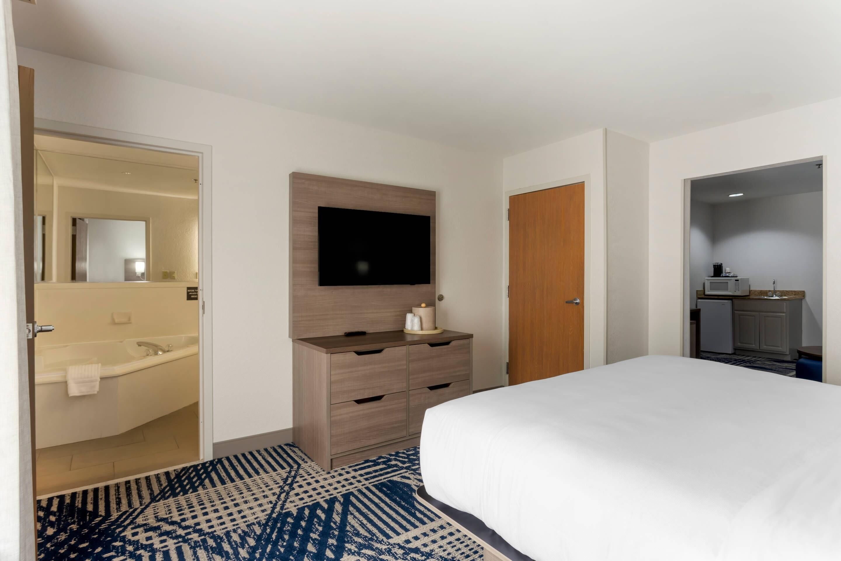 A hotel room with a bed, a TV mounted on the wall, a wooden dresser, a bathroom with a sink and mirror, and a kitchenette area.
