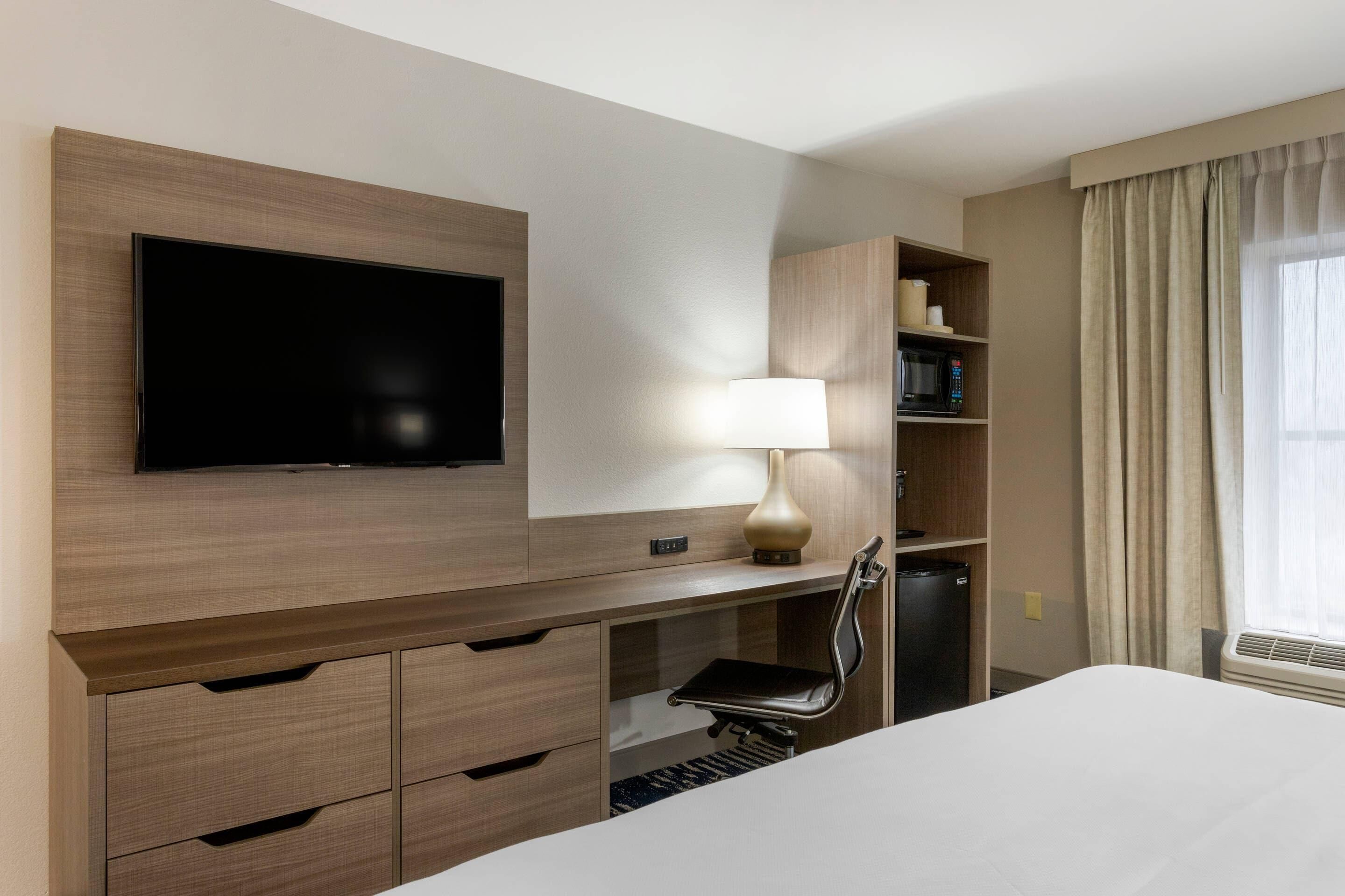 A hotel room with a flat-screen TV, a desk with a lamp, a chair, a bed, a mini-fridge, and a shelf with toiletries.