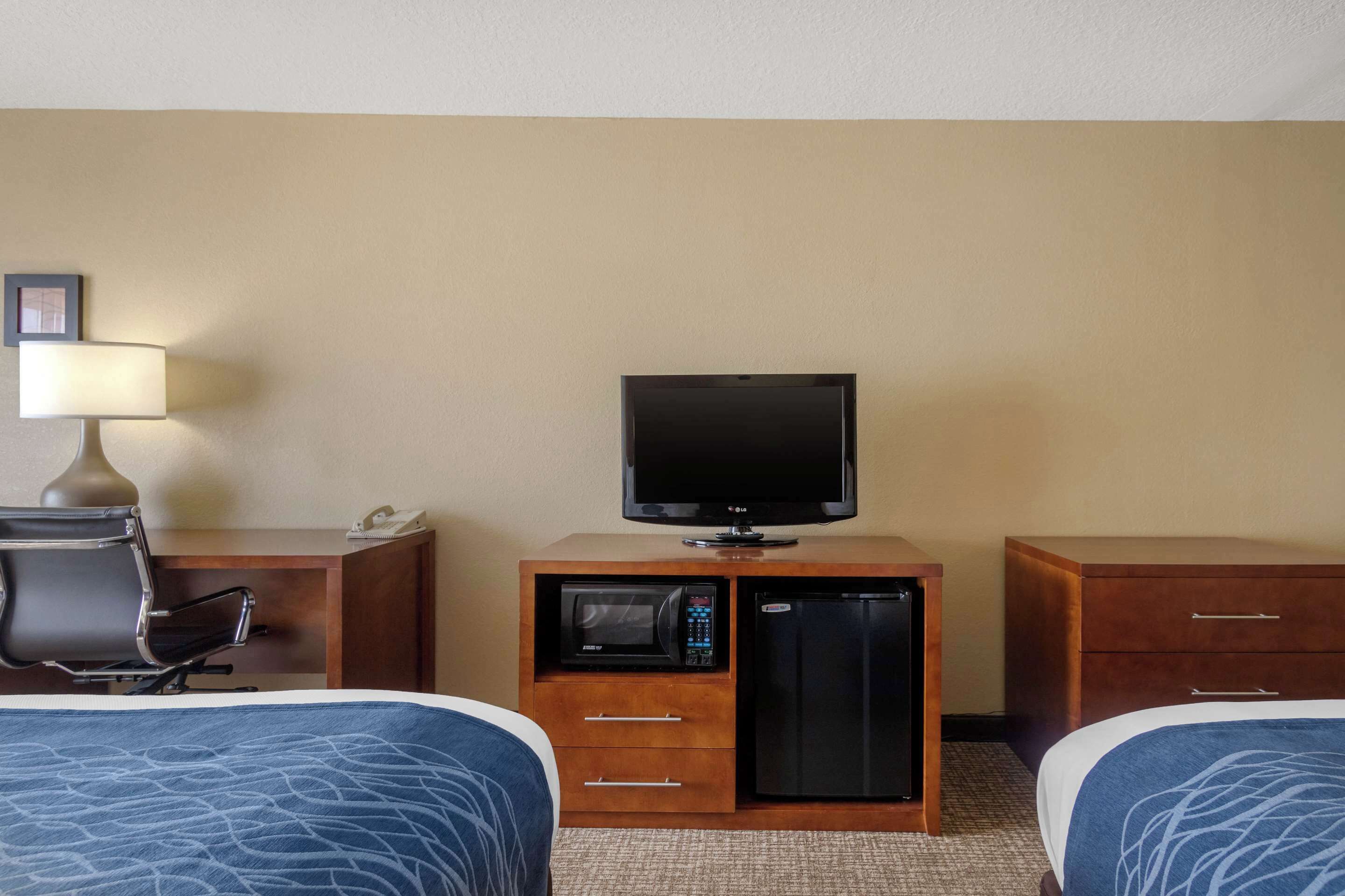 A hotel room with two beds, a television, a microwave, a refrigerator, a desk with a lamp, and a chair.