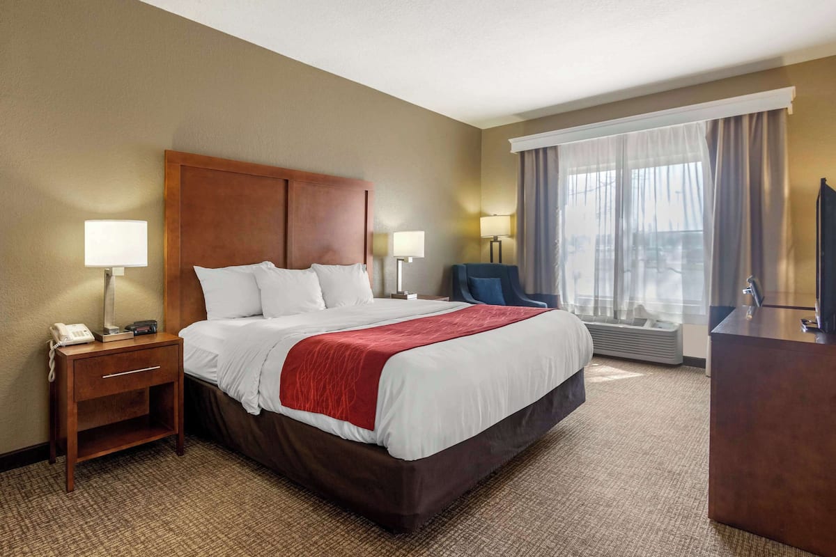 A hotel room with a large bed, two bedside tables with lamps, a desk with a chair, and a large window with curtains.