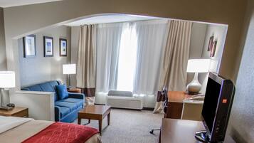 Une chambre d’hôtel avec un lit, un canapé bleu, un bureau avec un ordinateur, une lampe et une grande fenêtre avec des rideaux.