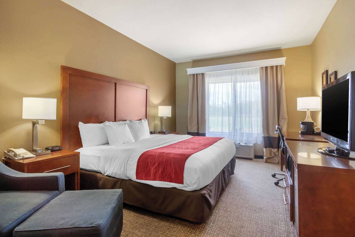 A hotel room with a large bed, a desk with a flat-screen TV, a chair, and a window with curtains.