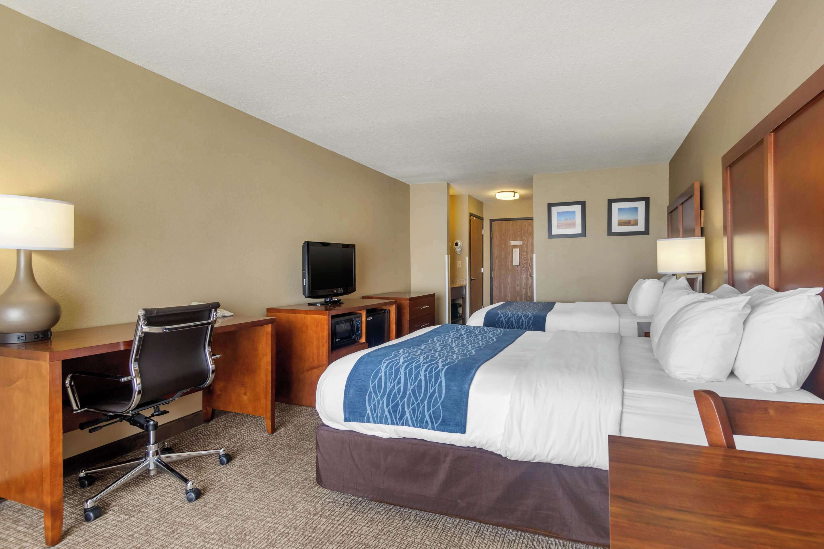 Quarto de hotel com cama, escrivaninha, cadeira, televisão e duas molduras na parede.