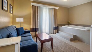 Habitación de hotel con sofá, una mesita, baño con bañera y ventana con cortinas.
