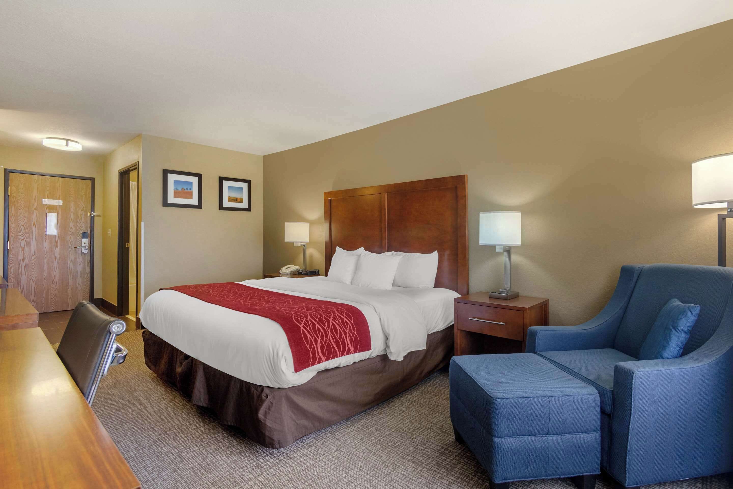 A hotel room with a large bed, a desk, a chair, and a nightstand with lamps.