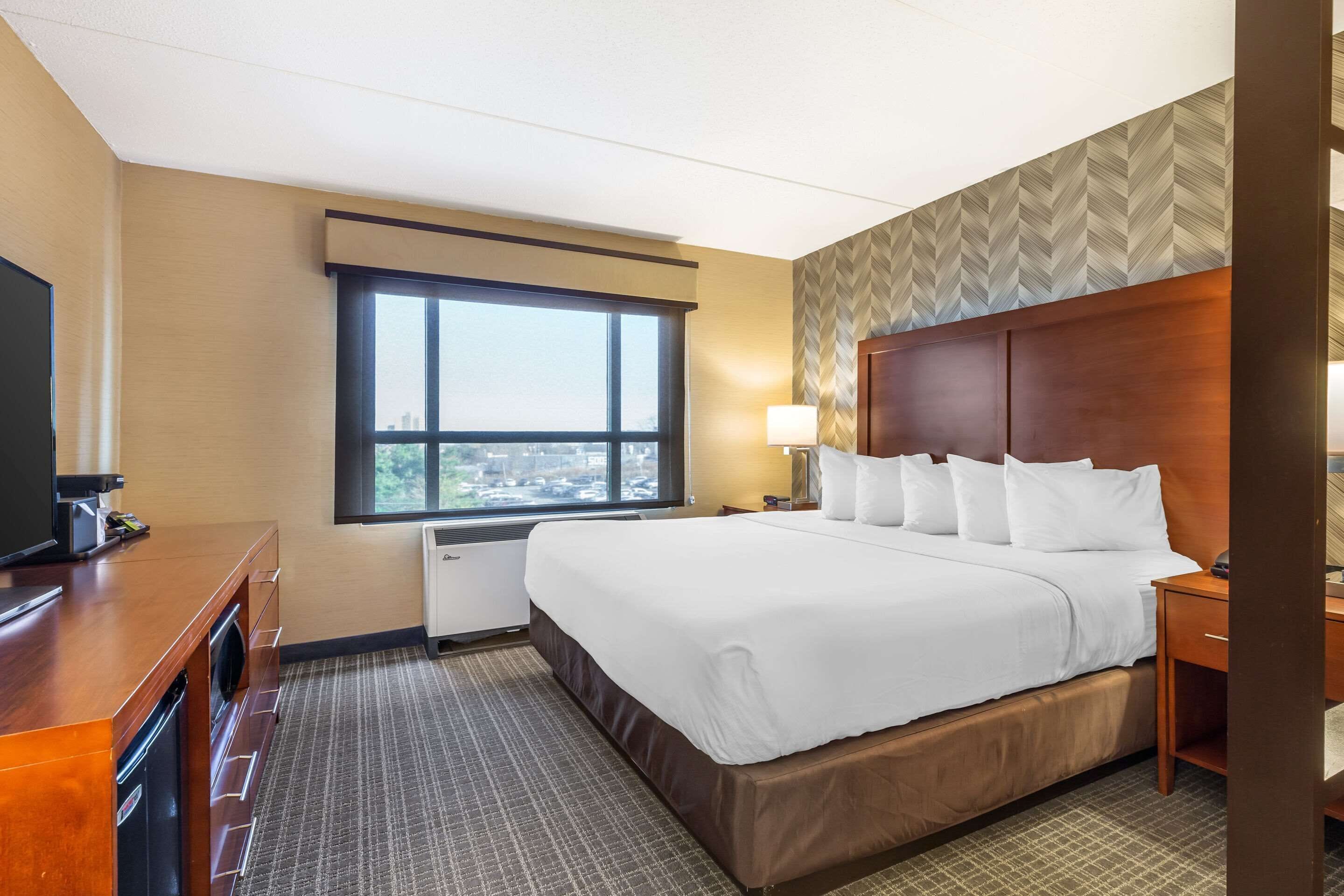 A hotel room with a large bed, a desk with a television, a window with a city view, and a patterned wall.