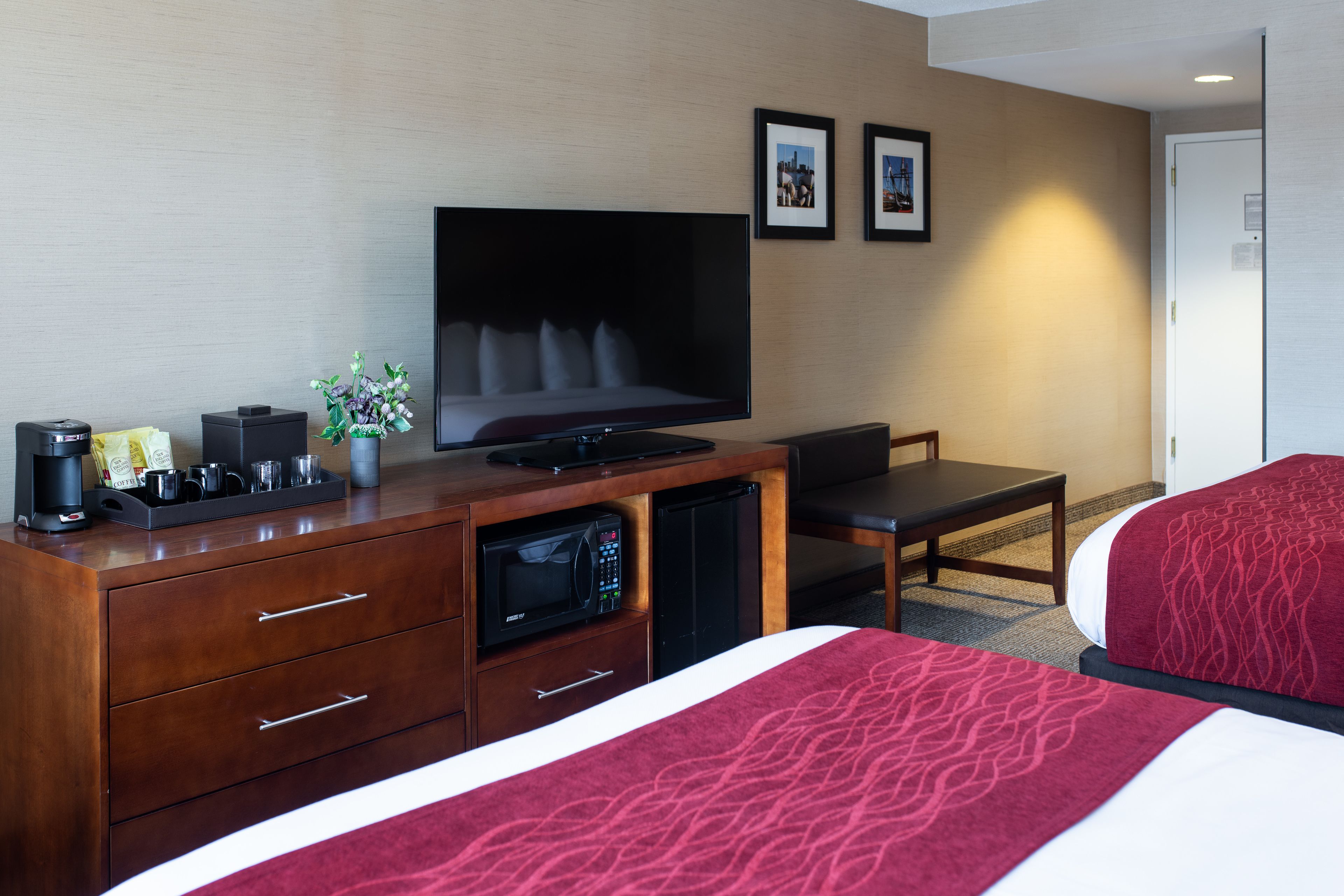 Una habitación de hotel con televisión, escritorio con cafetera, microondas, cama con manta roja y dos cuadros en la pared.