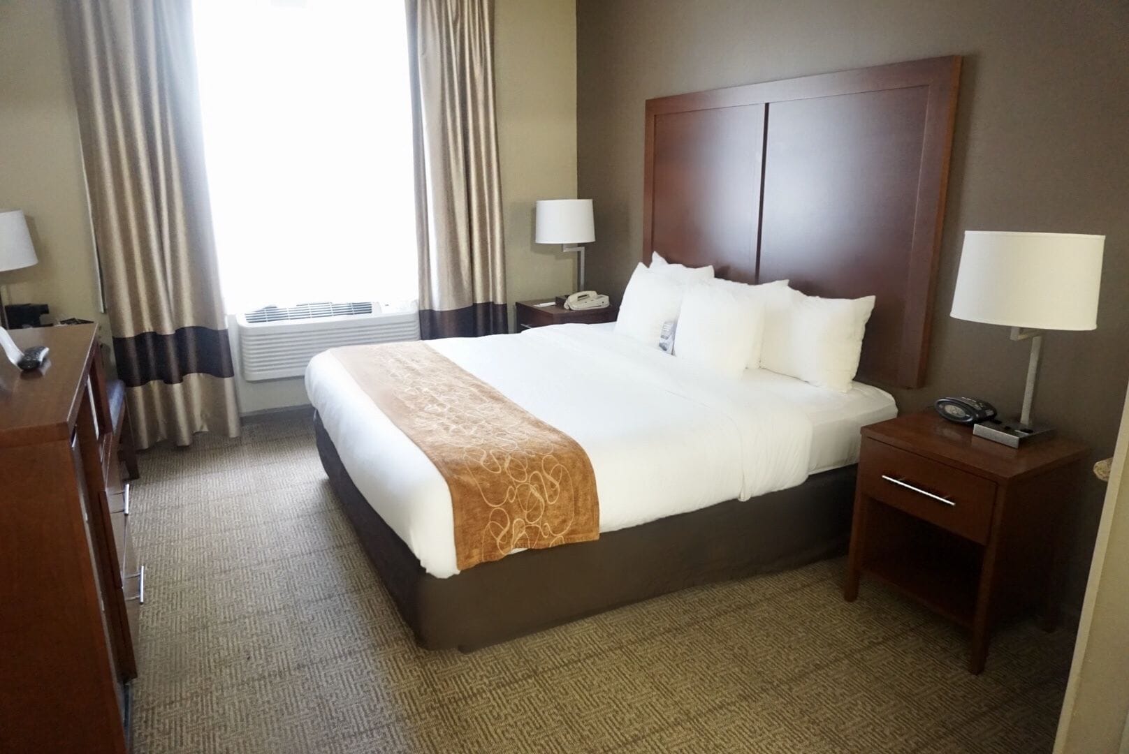 Habitación de hotel con una cama grande, un cabecero de madera, dos mesitas de noche con lámparas, un escritorio y una ventana con cortinas.