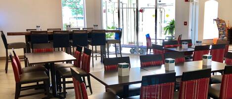 A spacious indoor dining area with multiple tables and chairs, large windows allowing natural light, and a view of the outside.