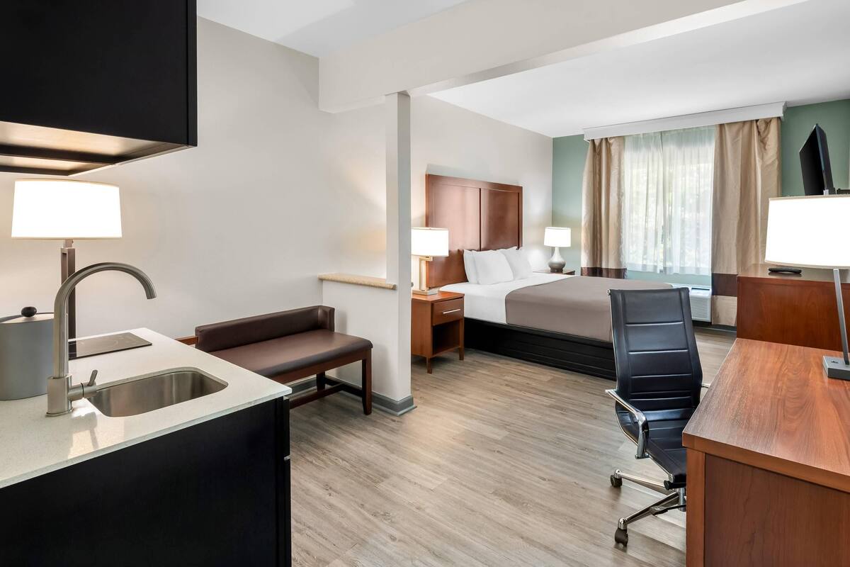 Quarto de hotel com cama, escrivaninha, cadeira e uma pequena área de cozinha.