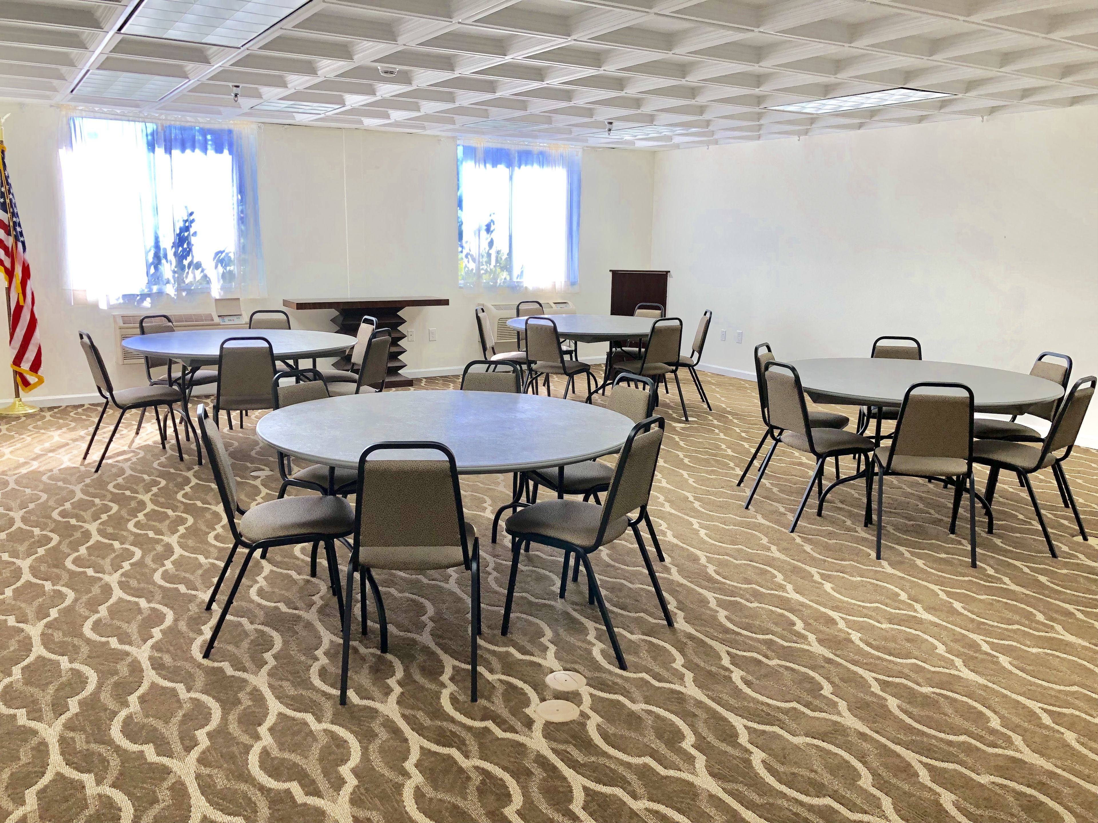 A conference room with multiple tables and chairs, arranged in a U-shape. There are two large windows with blinds, an American flag, and a ceiling with a grid pattern.