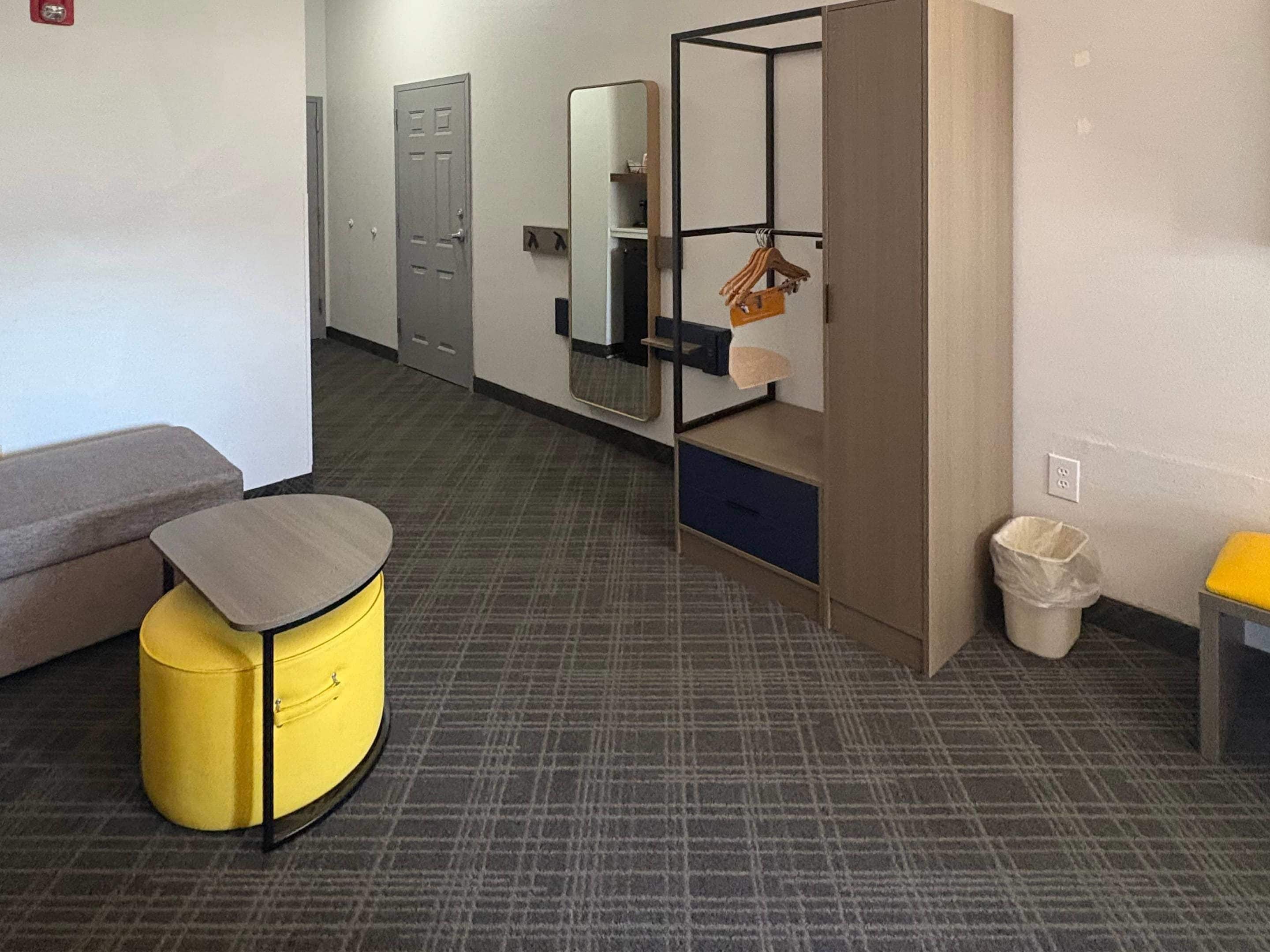 Una habitación con un sofá gris, un otomano amarillo, un espejo, un estante pequeño con un objeto colgando y un cubo de basura.