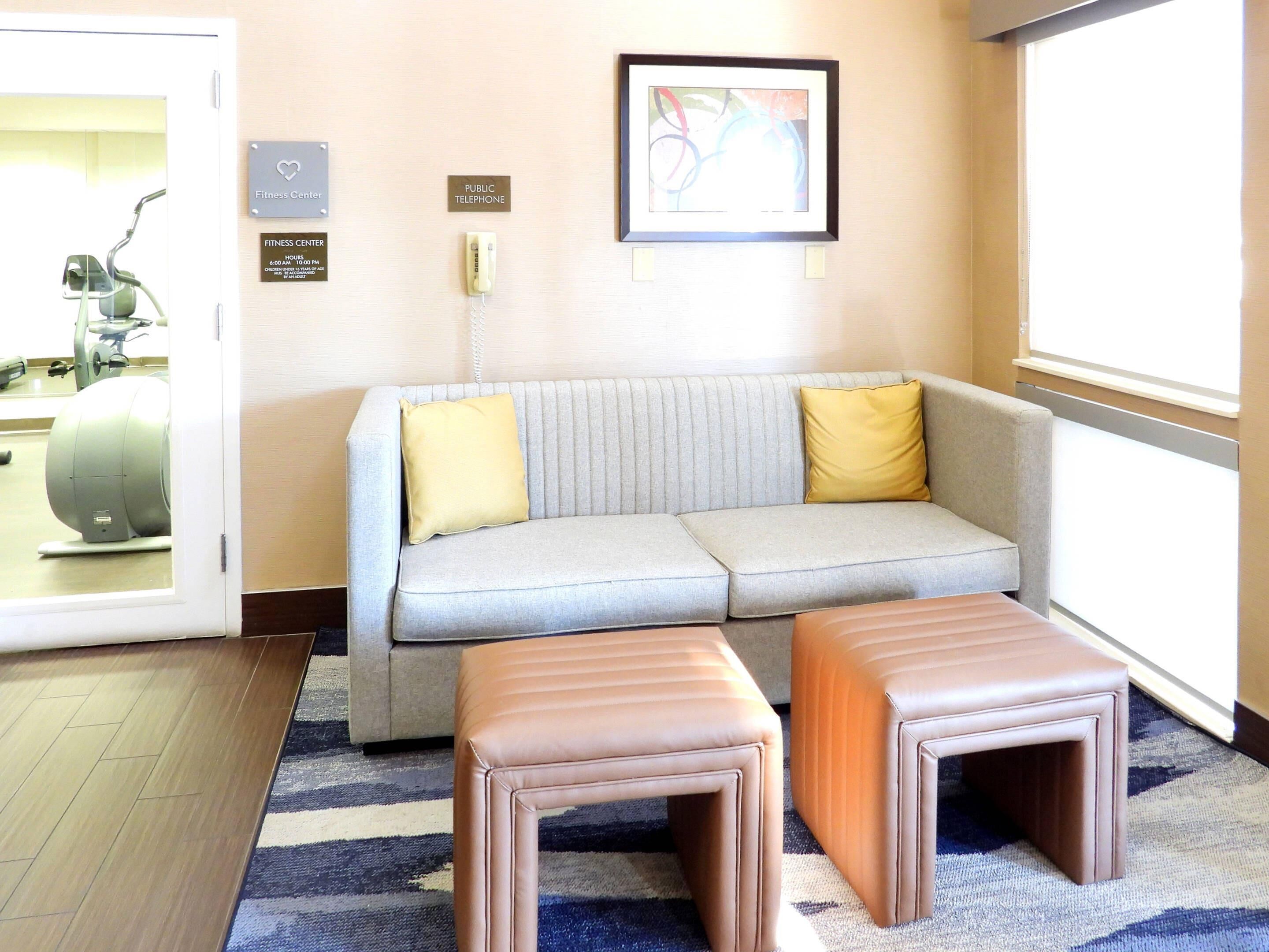 A reception area with a grey sofa, two brown ottomans, a fitness center sign, a public telephone, and a framed abstract artwork on the wall.
