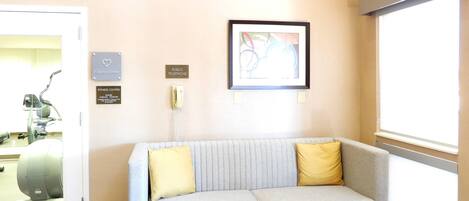 A reception area with a grey sofa, two brown ottomans, a fitness center sign, a public telephone, and a framed abstract artwork on the wall.