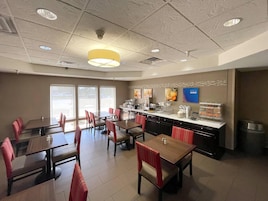 A restaurant with multiple tables and chairs, a counter with food items, and a large window providing a view outside.