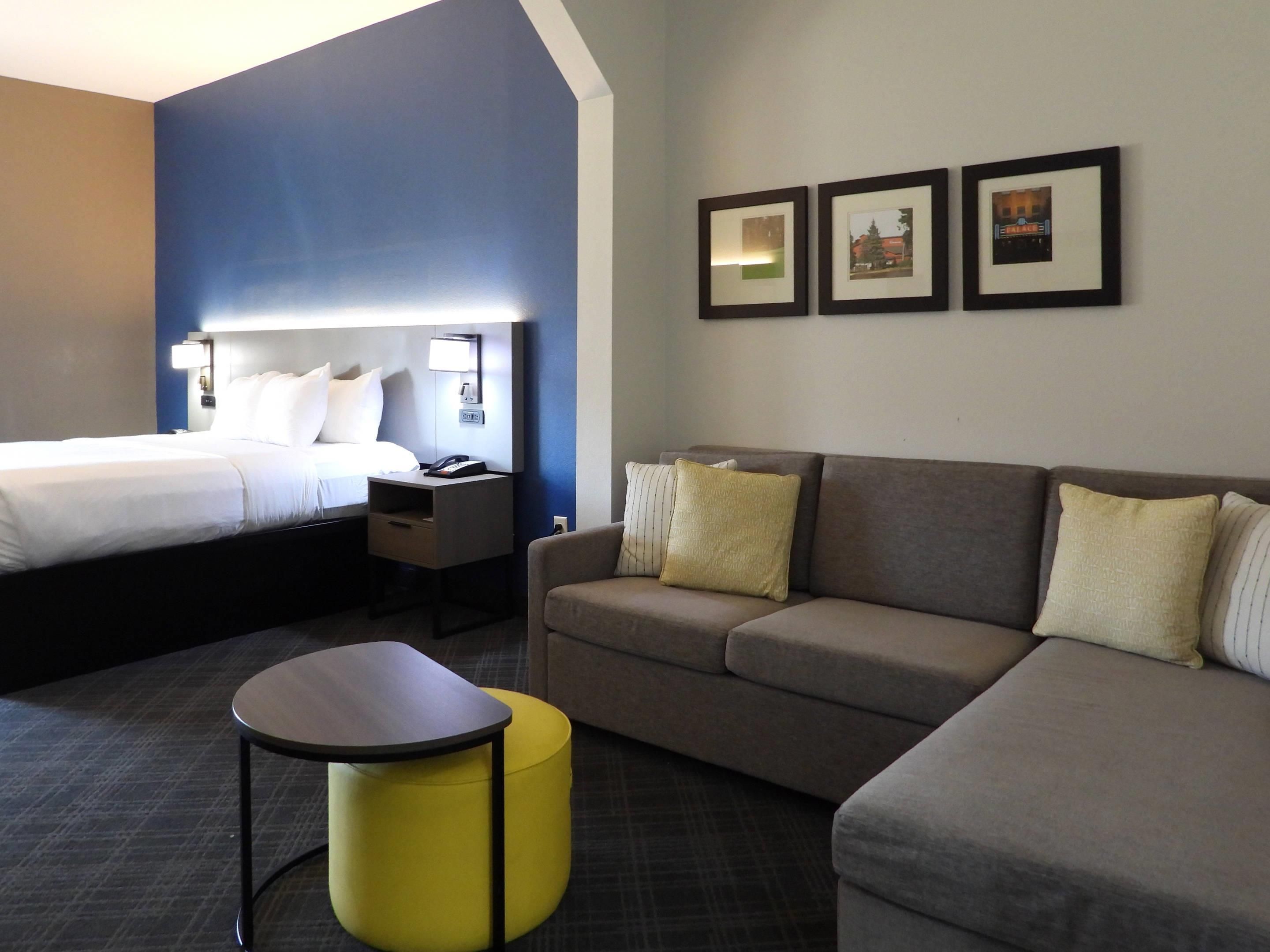 A hotel room with a bed, a sofa, a small table, and a yellow ottoman. There are framed pictures on the wall.