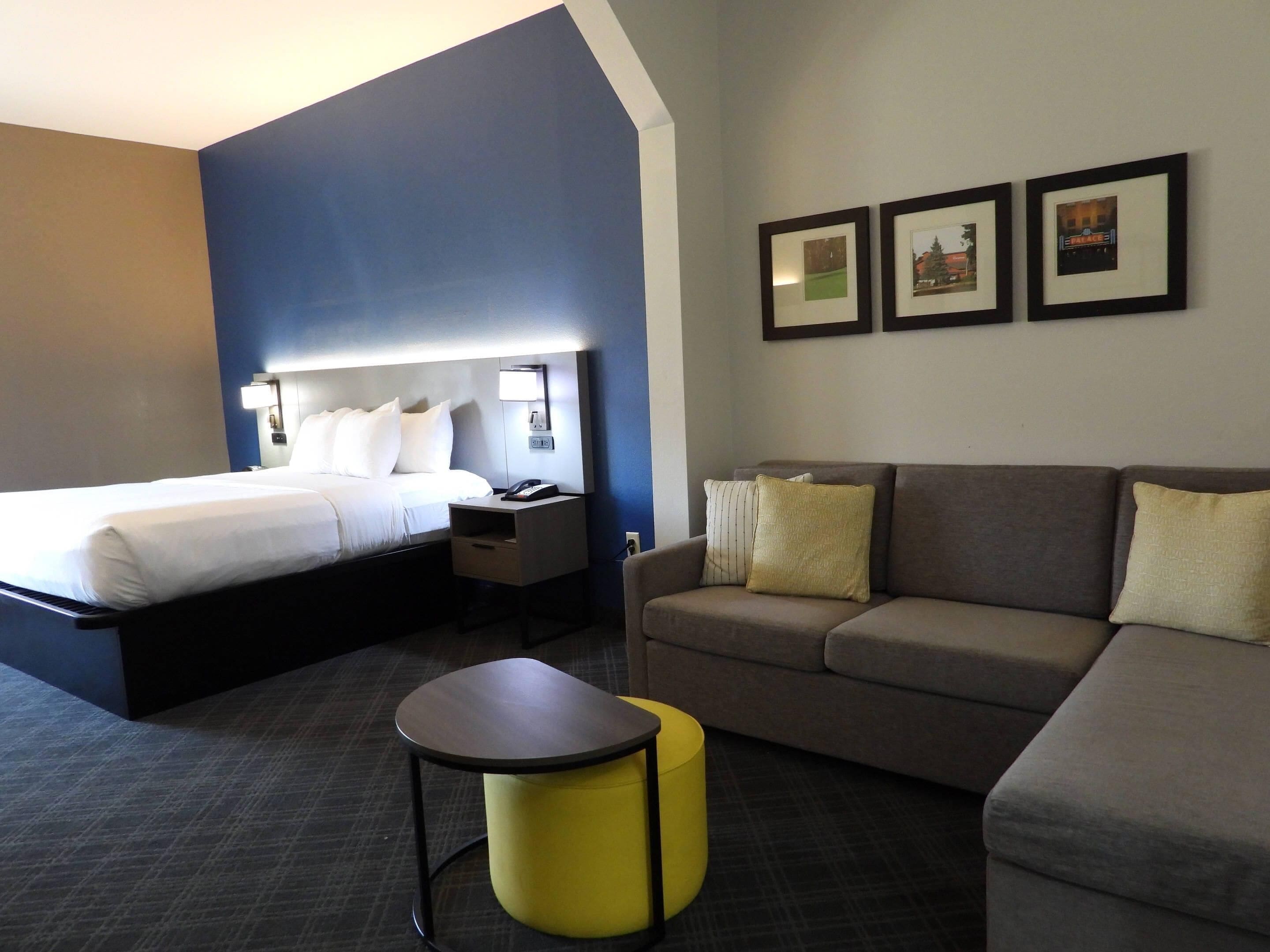 Habitación de hotel con una cama, un sofá, una mesita y una mesita de noche. En la pared hay cuadros enmarcados.